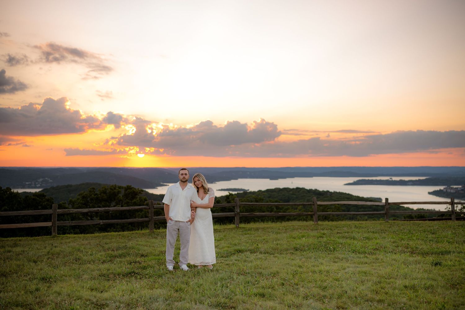Couple in white attire poses on green hillside at golden sunset overlooking a lake.