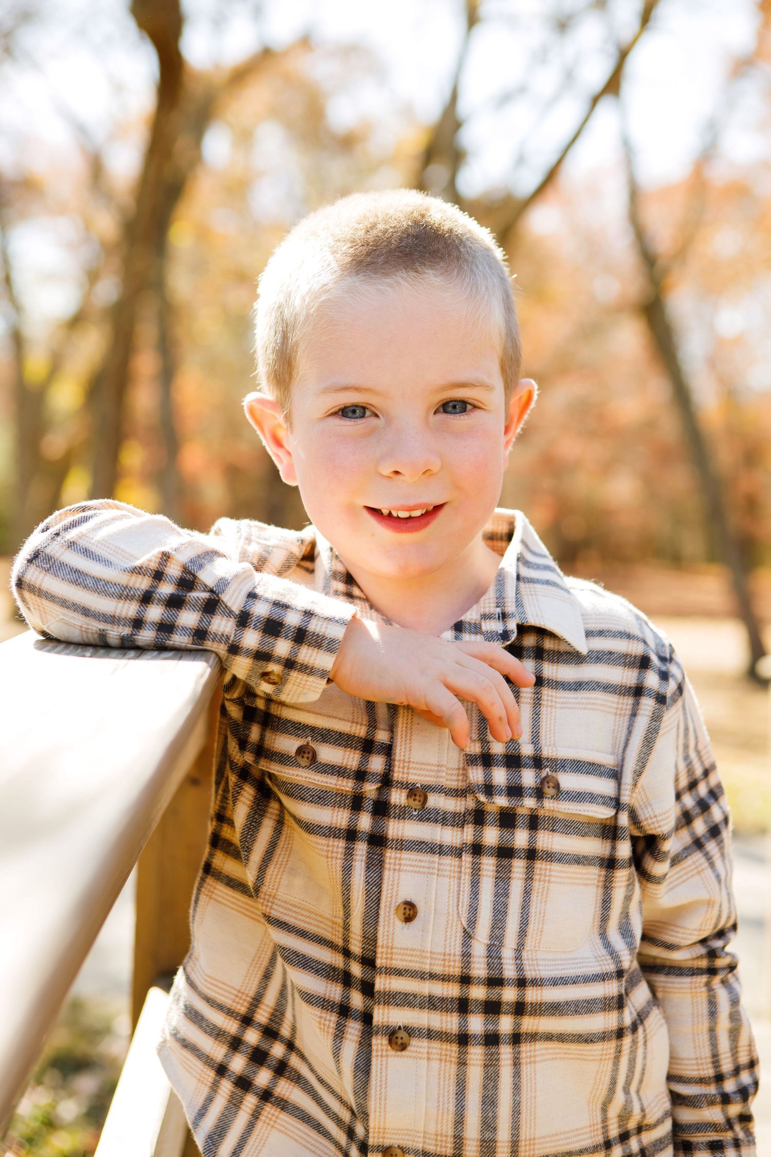 Young boy in plaid shirt smiling while leaning on wooden bridge railing in fall park.