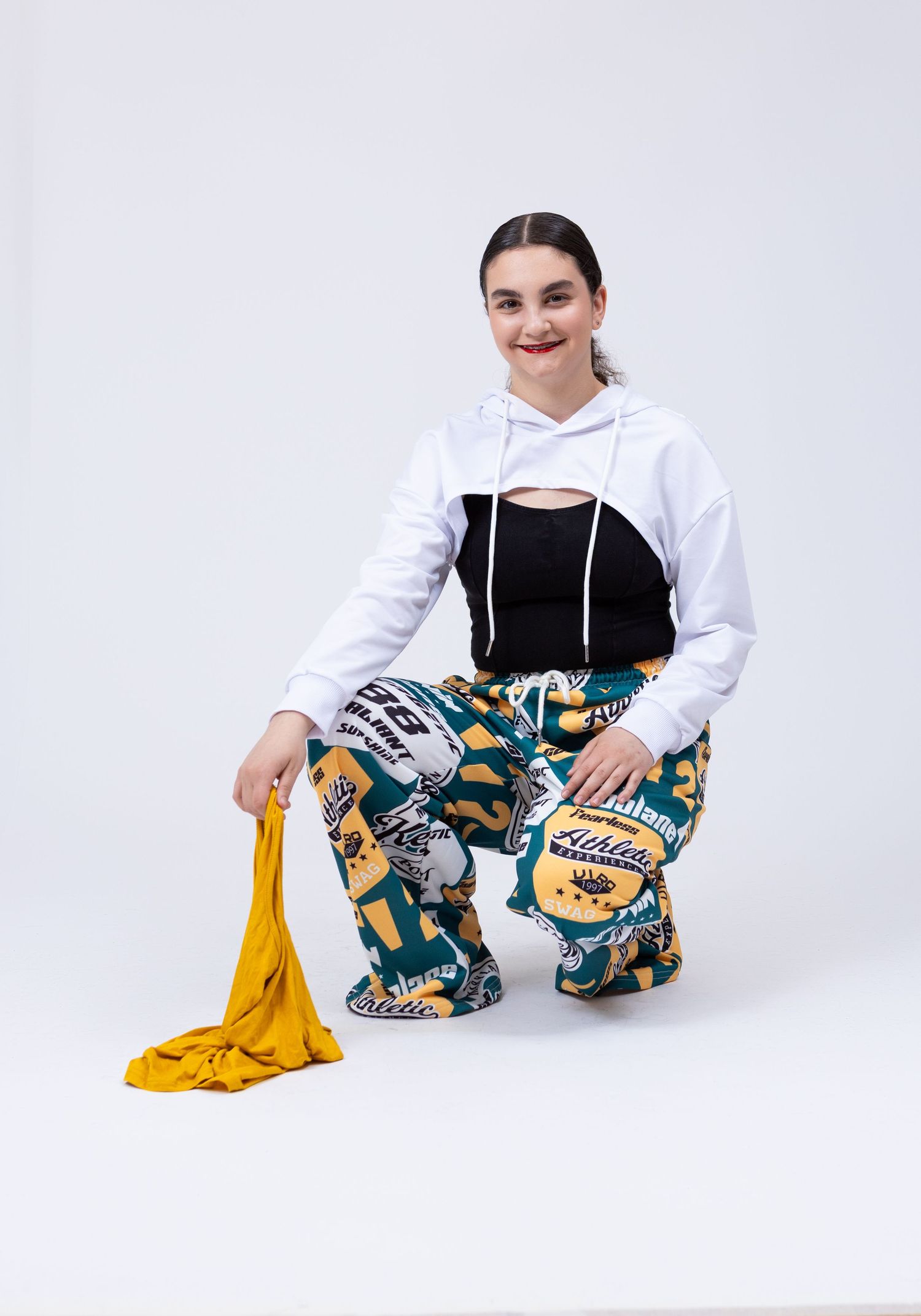 Young woman in white crop hoodie and colorful printed pants crouches holding yellow fabric.