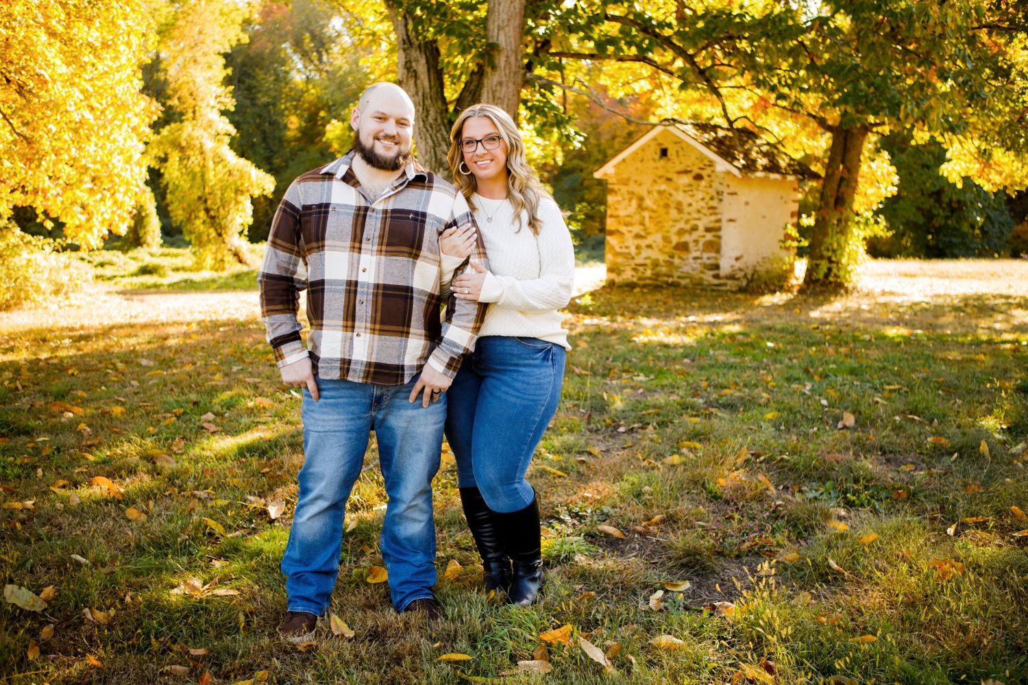 Smiling couple standing together in a sunny autumn park with golden foliage behind them.