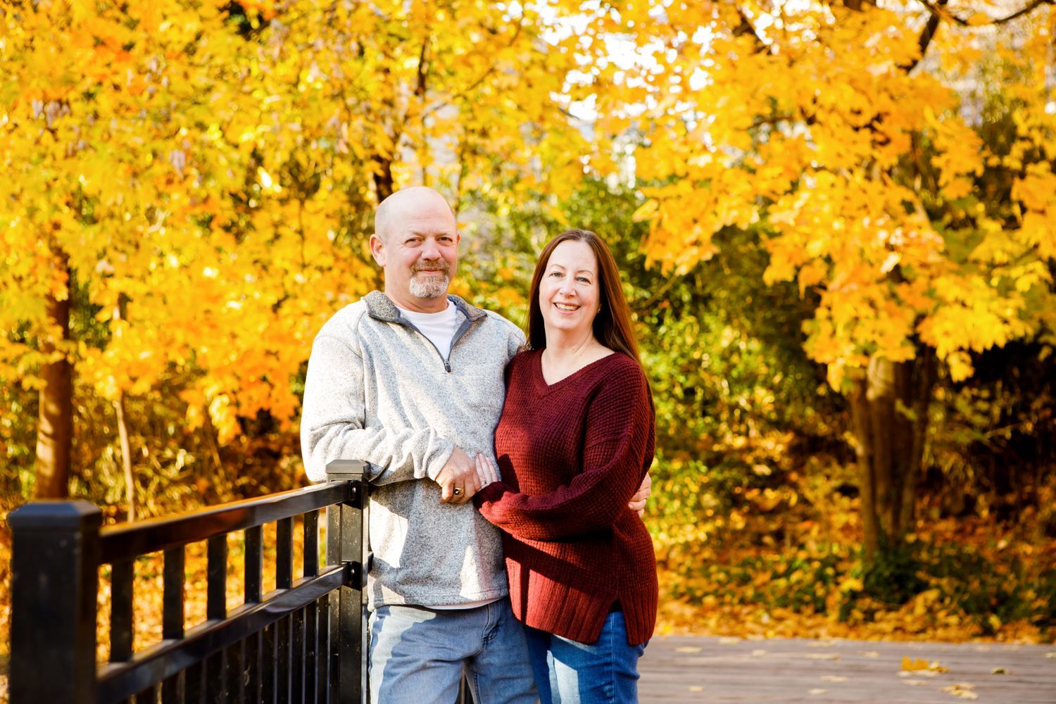 Couple smiling together on a wooden bridge surrounded by vibrant yellow autumn foliage.