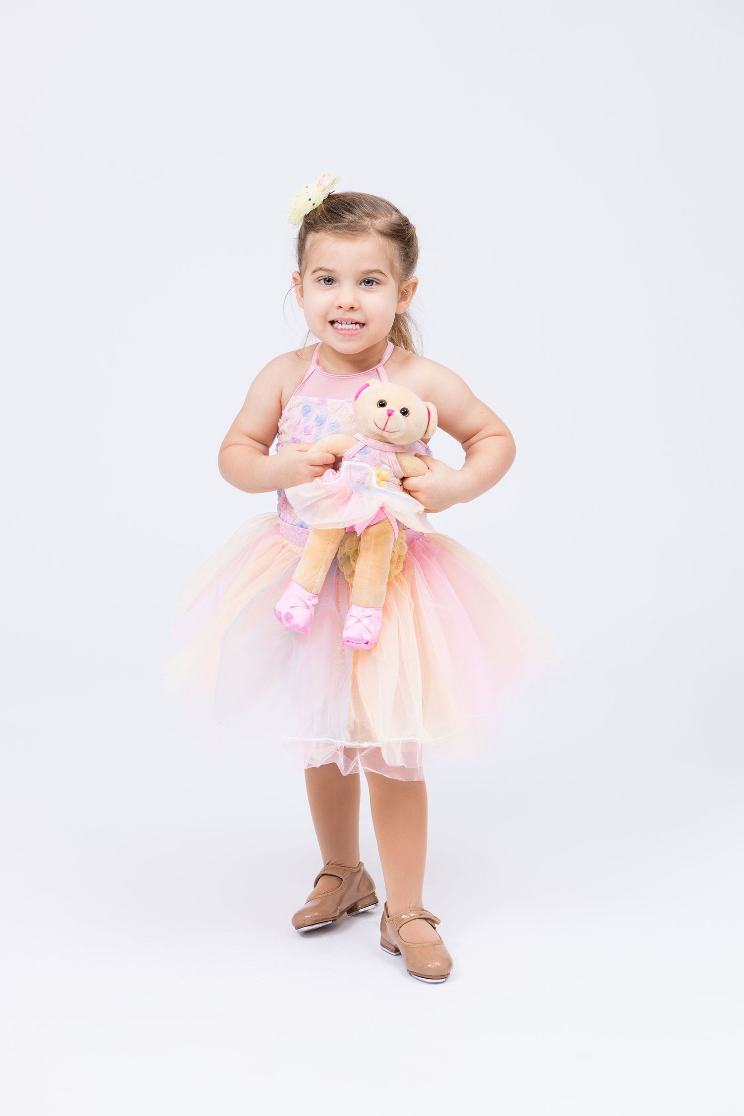 Adorable young girl in pink tutu dress smiles while holding a stuffed ballerina doll toy.