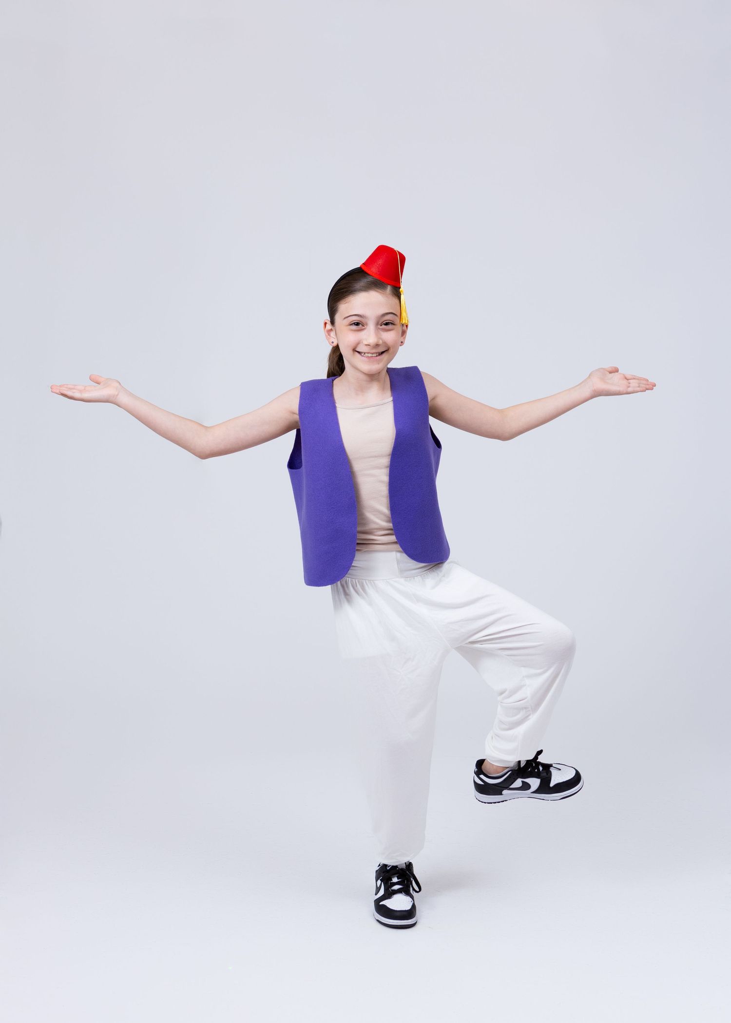 Child dancer in purple vest and red fez hat balances on one leg with arms spread wide.