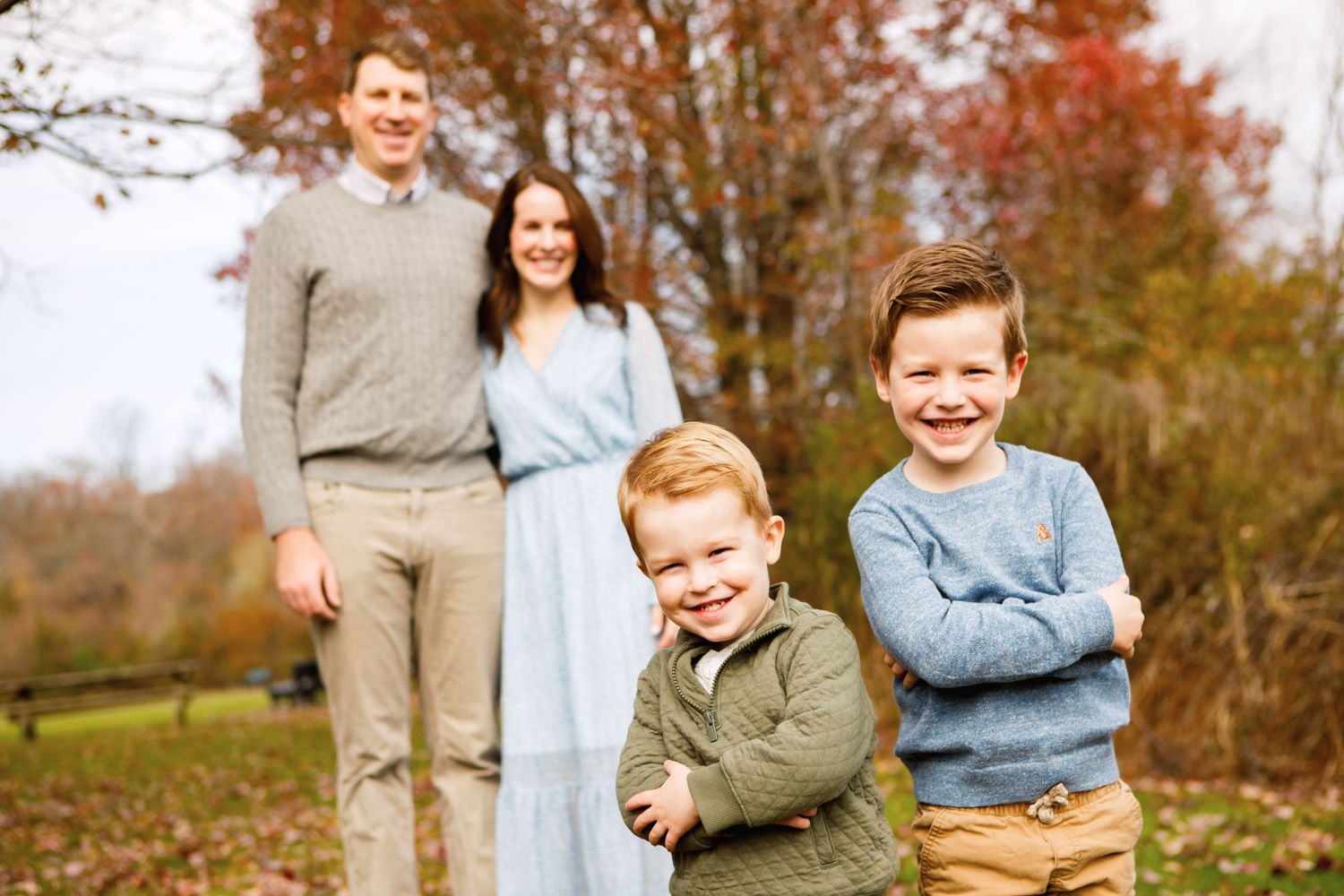 Family of four poses in an autumn park, two young boys in front with parents standing behind.