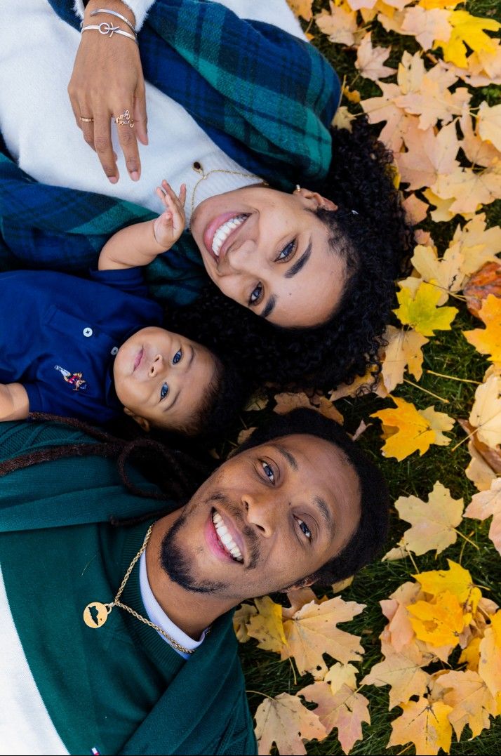 Family of three lies in autumn leaves smiling, parents with young toddler between them overhead view.