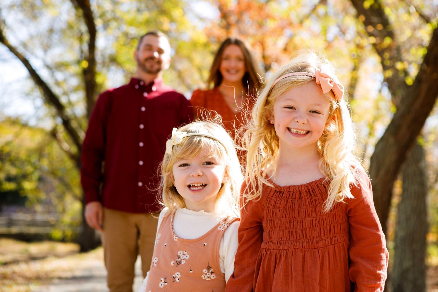 Two blonde sisters smiling with parents in a sunny autumn park.