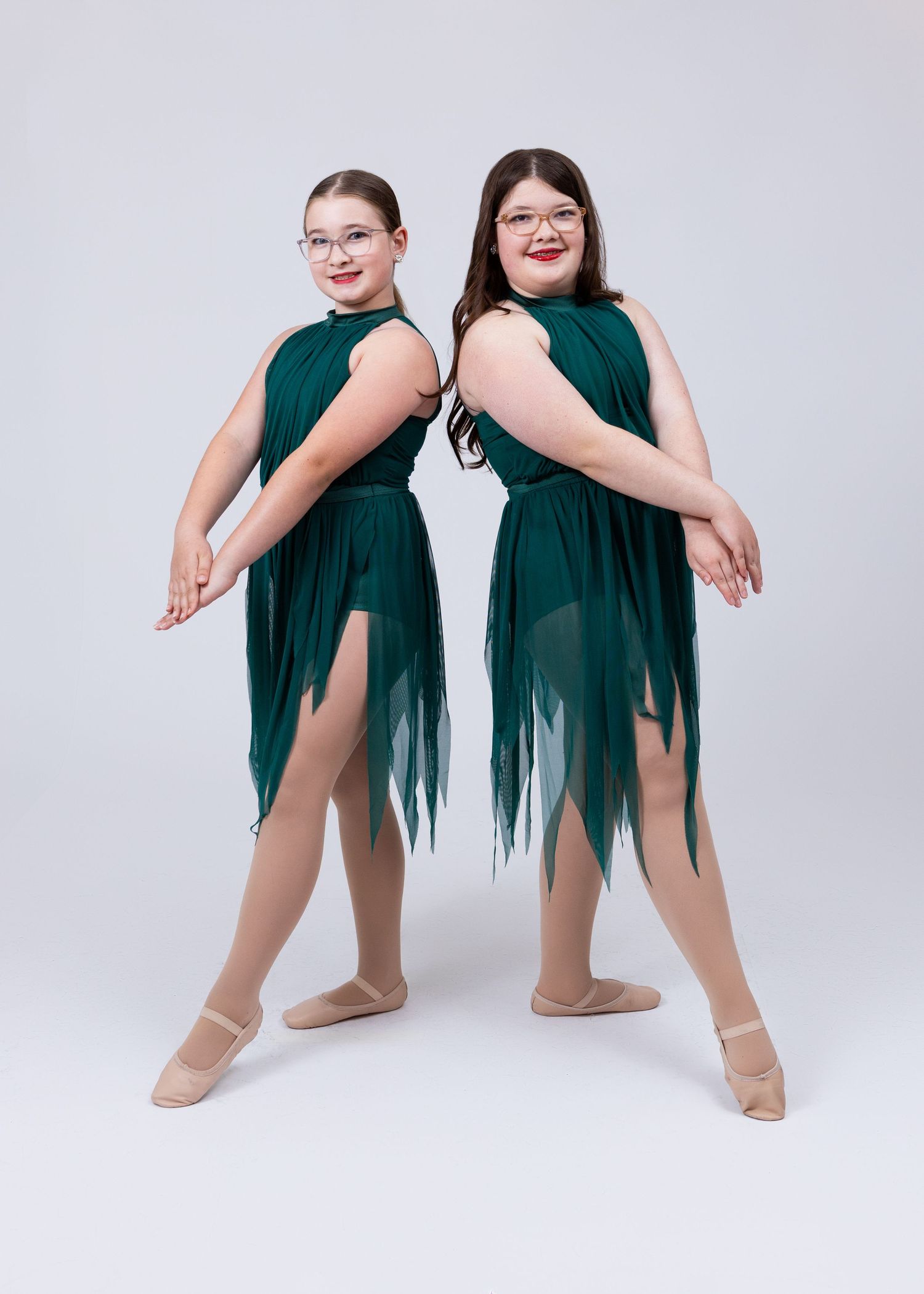 Two young dancers in matching dark green asymmetrical dresses pose back-to-back in ballet shoes.