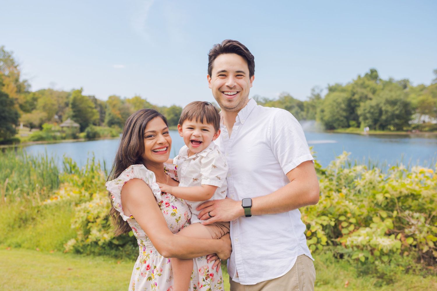 A happy family with a toddler poses outdoors near a serene lake on a sunny day.