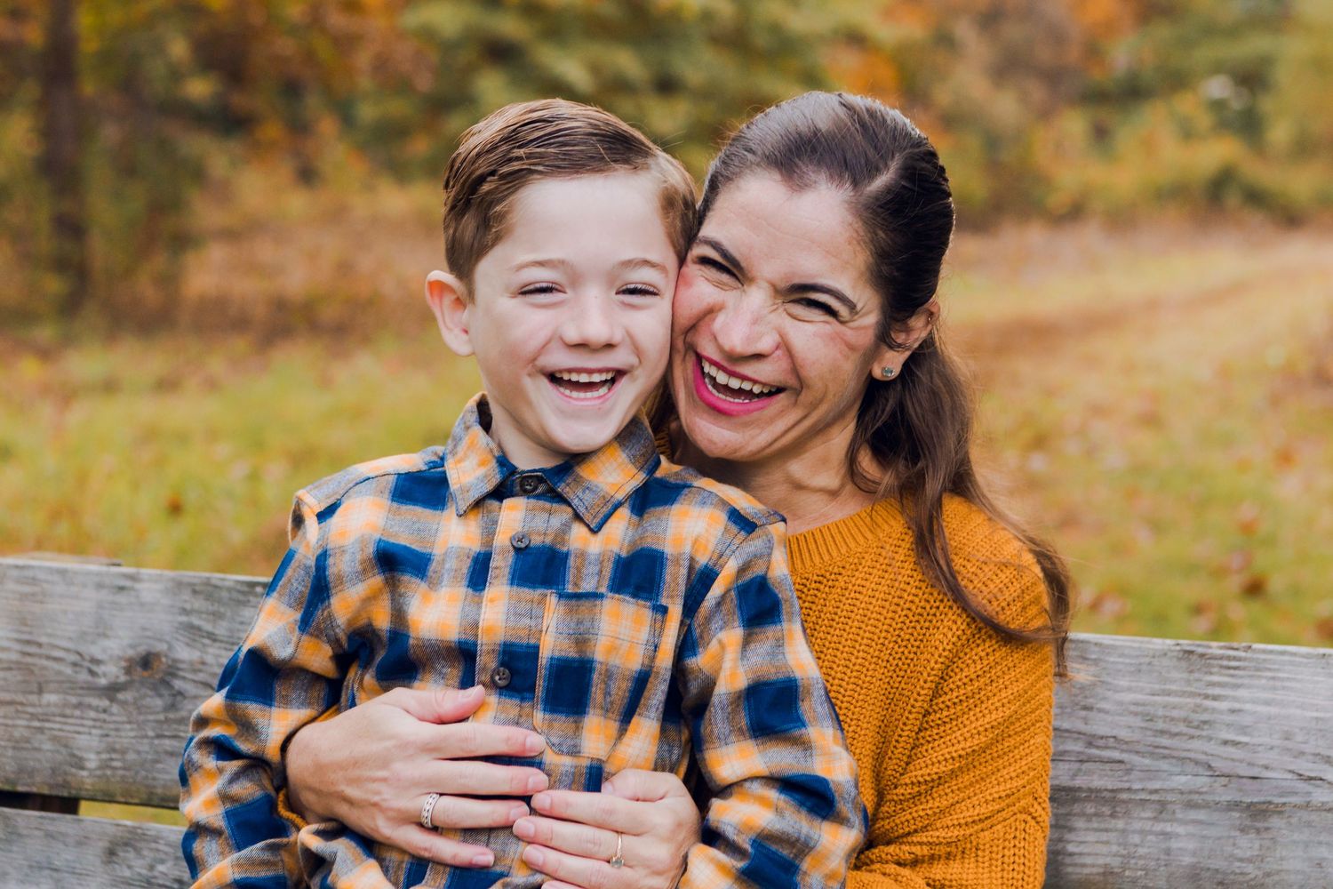 A woman in an orange sweater hugs a smiling boy in a plaid shirt on a bench with fall foliage backdrop.