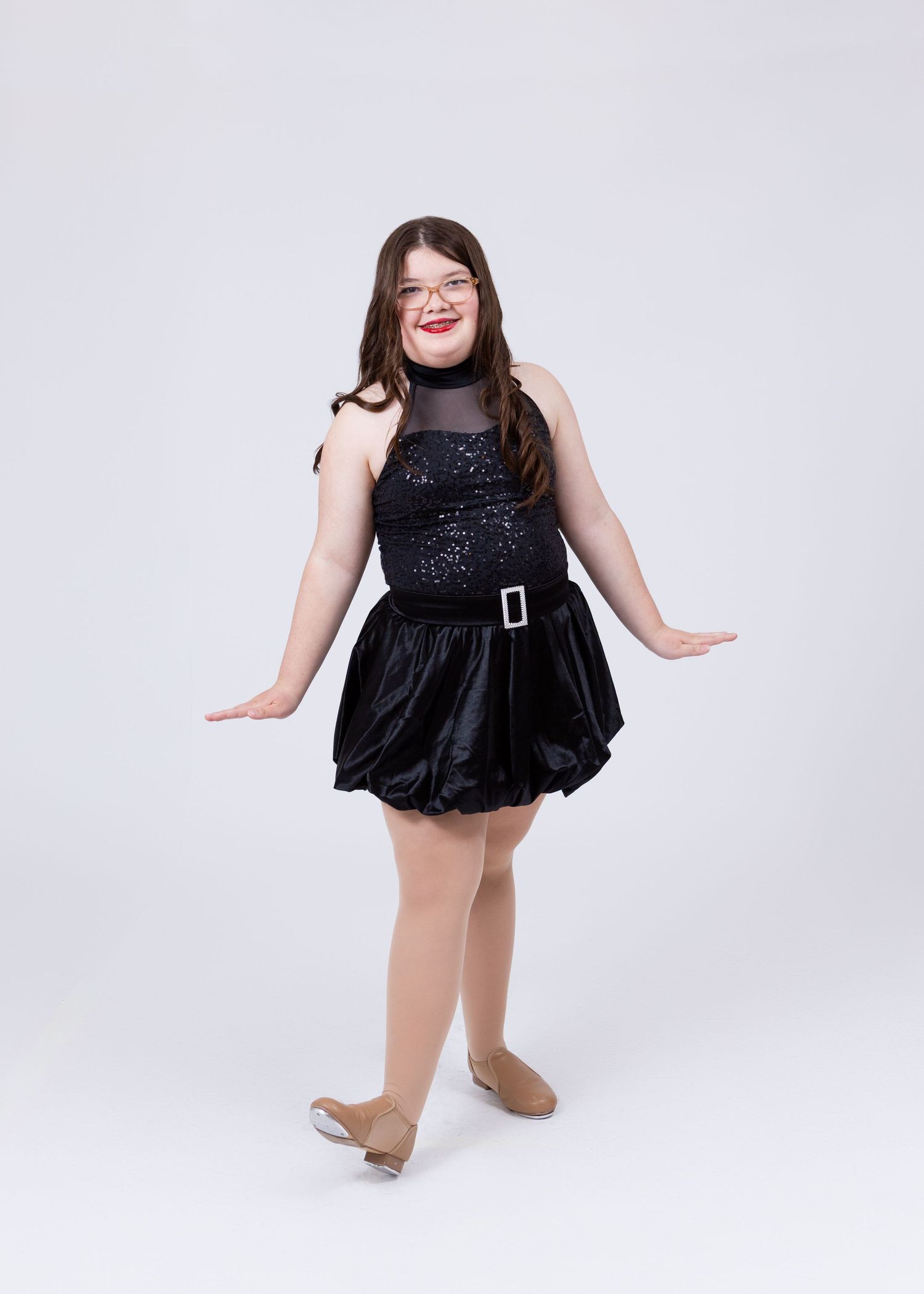 A young dancer in a black sequin bubble skirt costume poses with arms extended outward.
