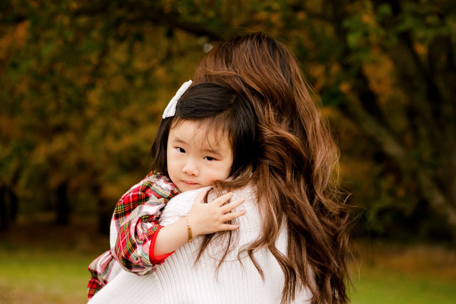 Young toddler girl in plaid scarf hugs mother with long brown hair in outdoor autumn setting.