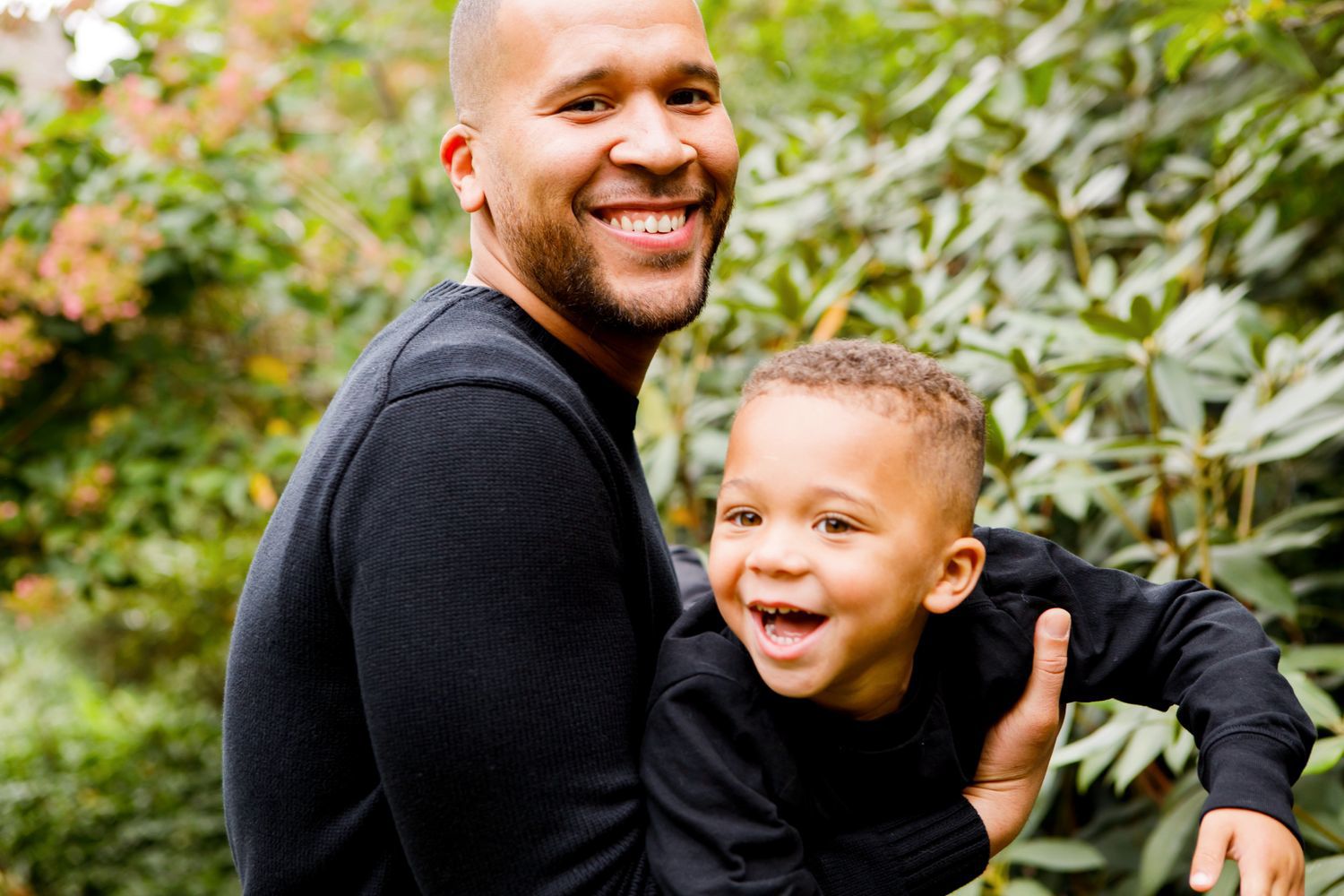Smiling bald man holds a laughing young boy outdoors surrounded by green foliage.
