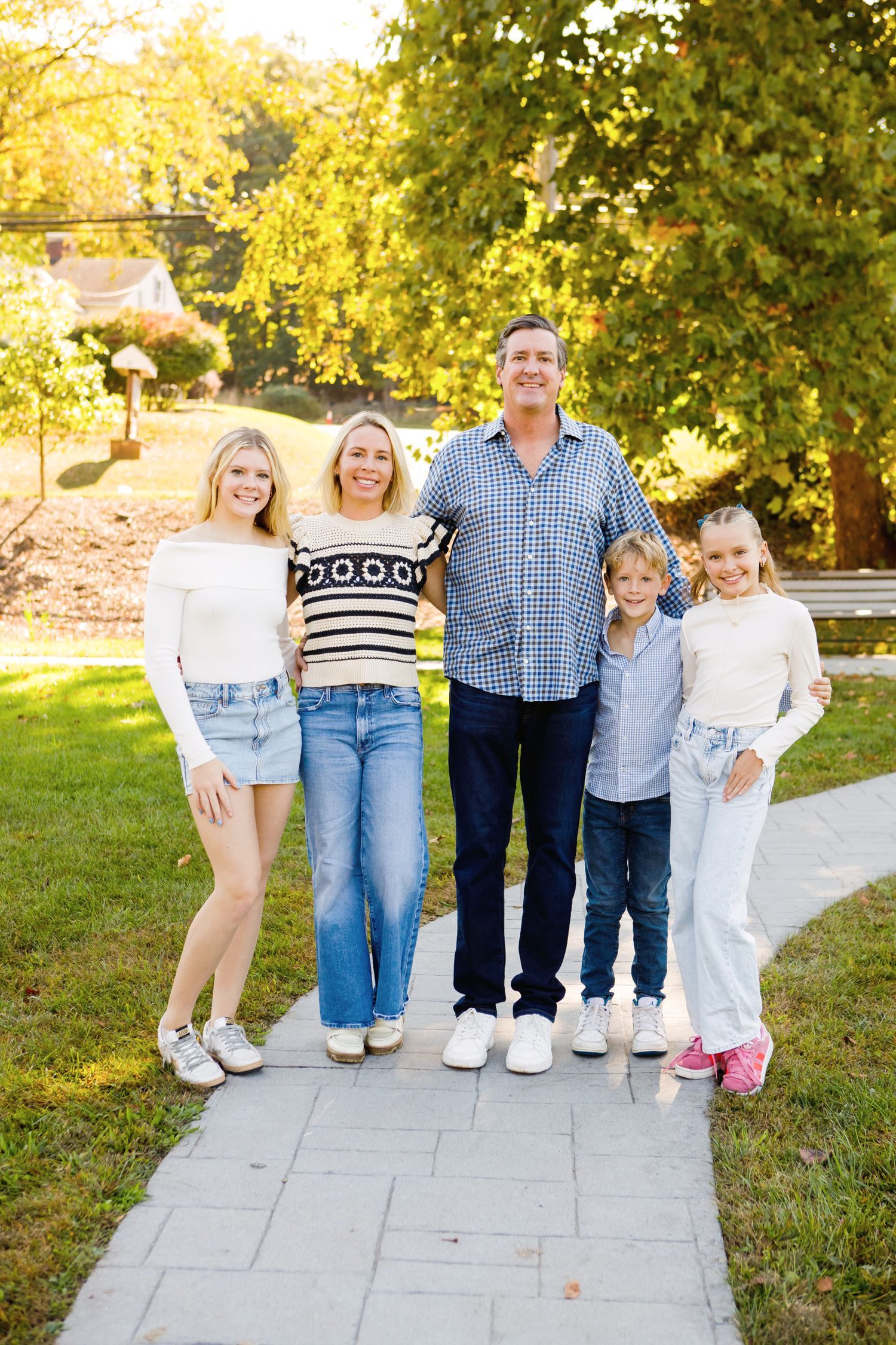 Family of five smiling outdoors on a stone path surrounded by green trees in warm sunlight.