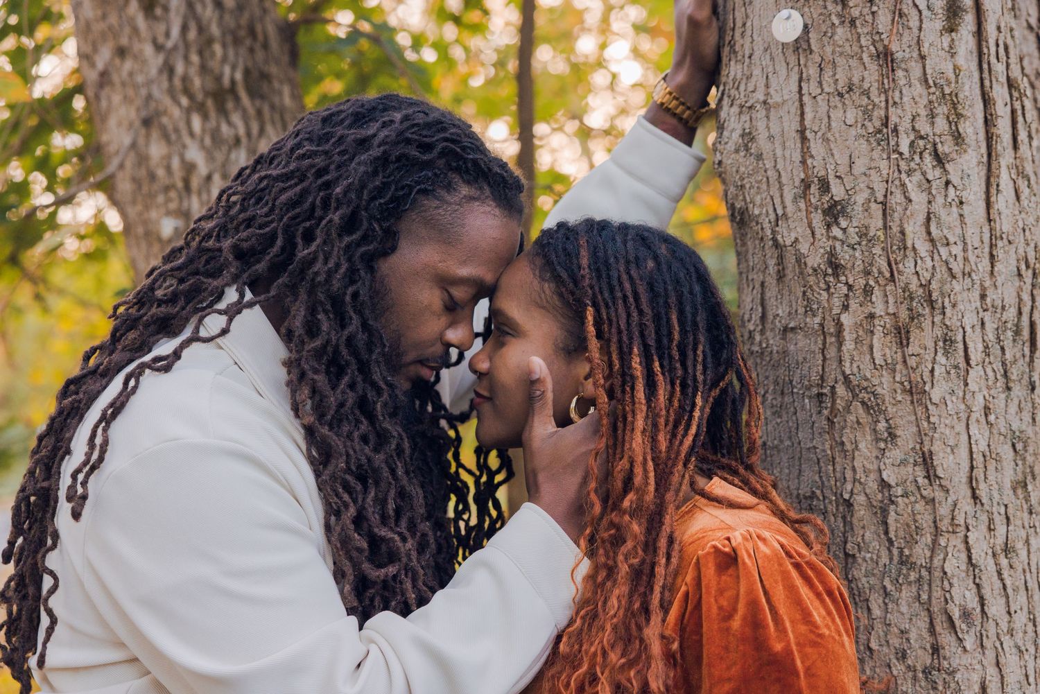 Romantic couple with dreadlocks sharing an intimate moment leaning against a tree in fall park.