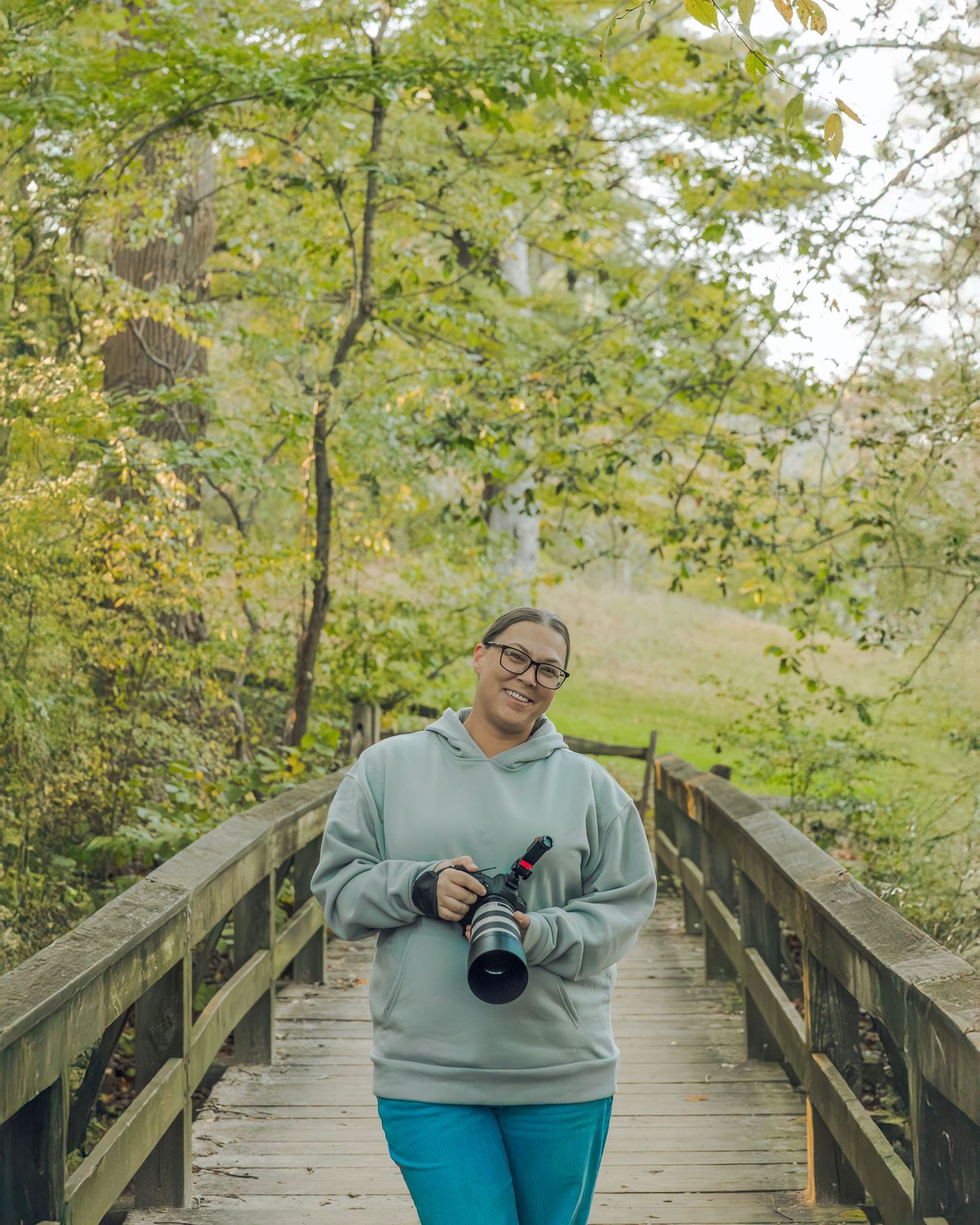 Smiling female photographer holding camera on wooden bridge surrounded by lush green trees.