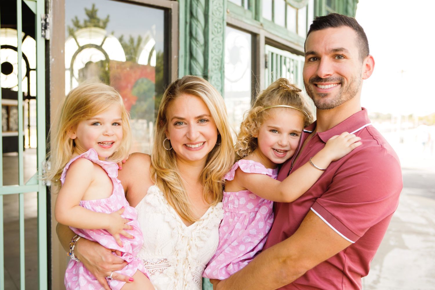 Happy family of four posing outdoors, parents holding two young blonde girls in matching pink dresses.