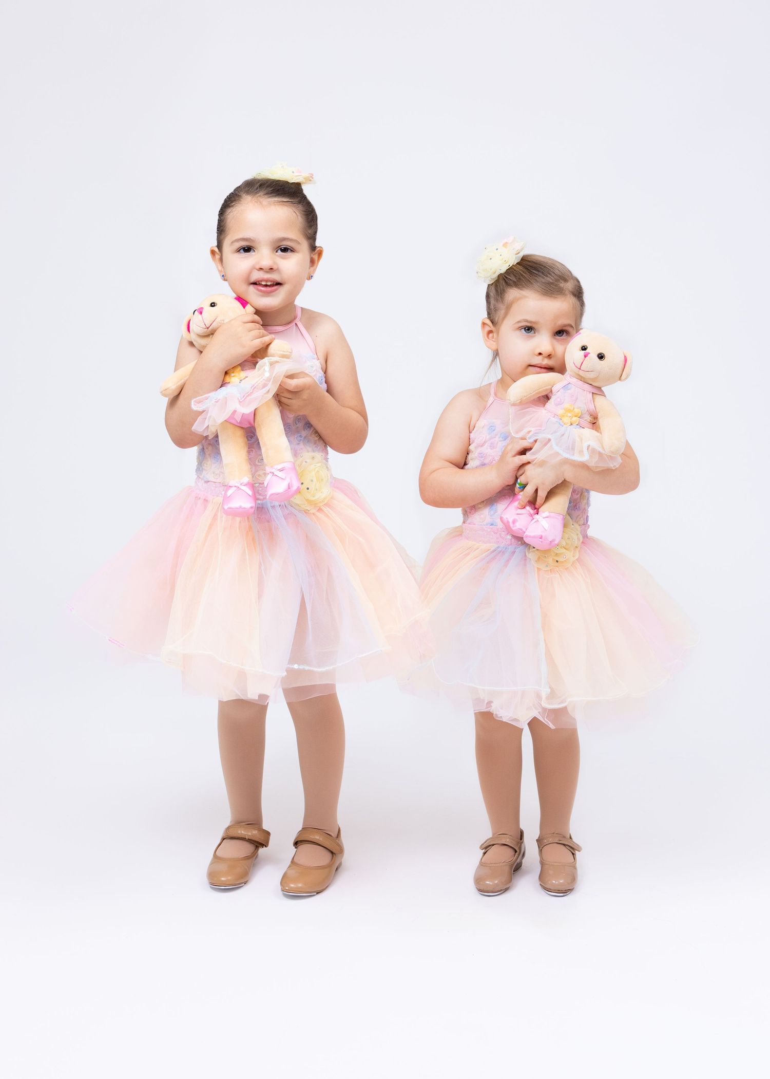 Two toddler girls in pink tutu dresses hold stuffed bear dolls in matching ballet costumes.