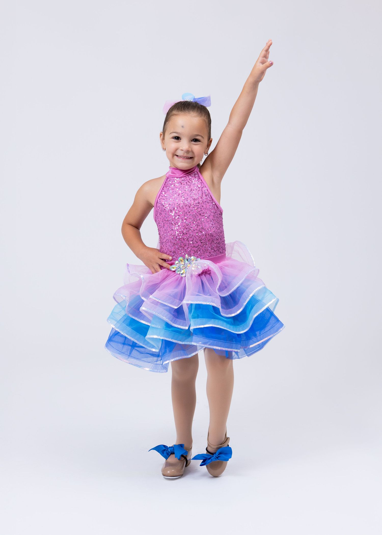 Young girl in pink sequin and rainbow tutu dance costume poses with arm raised in studio.