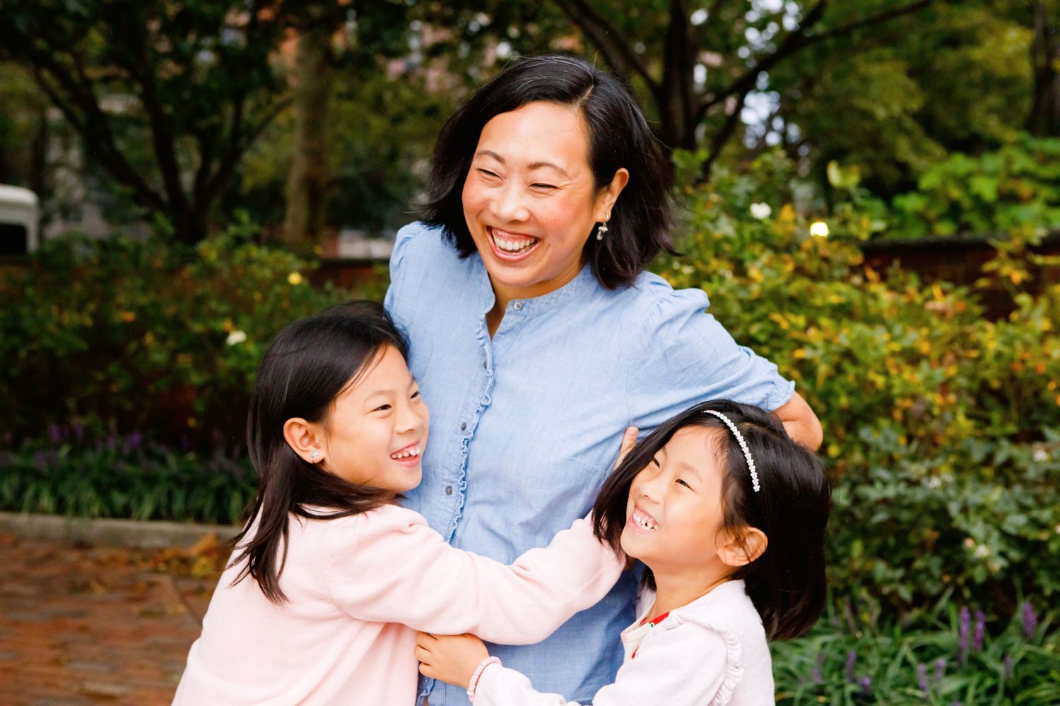 Smiling mother hugging her two young daughters in a lush garden setting.