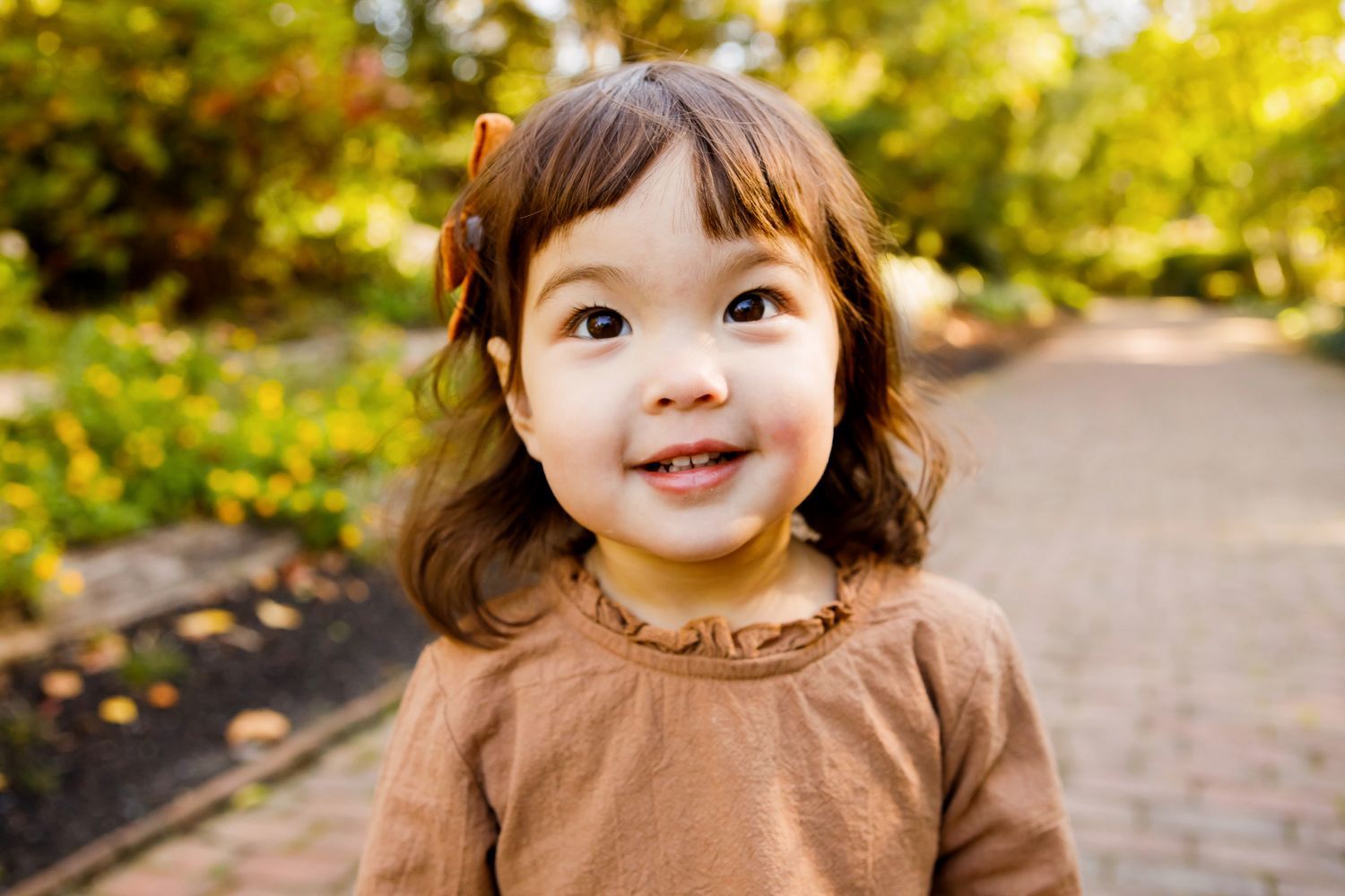 Adorable toddler girl with brown hair and a hair clip smiles on a sunny garden pathway.