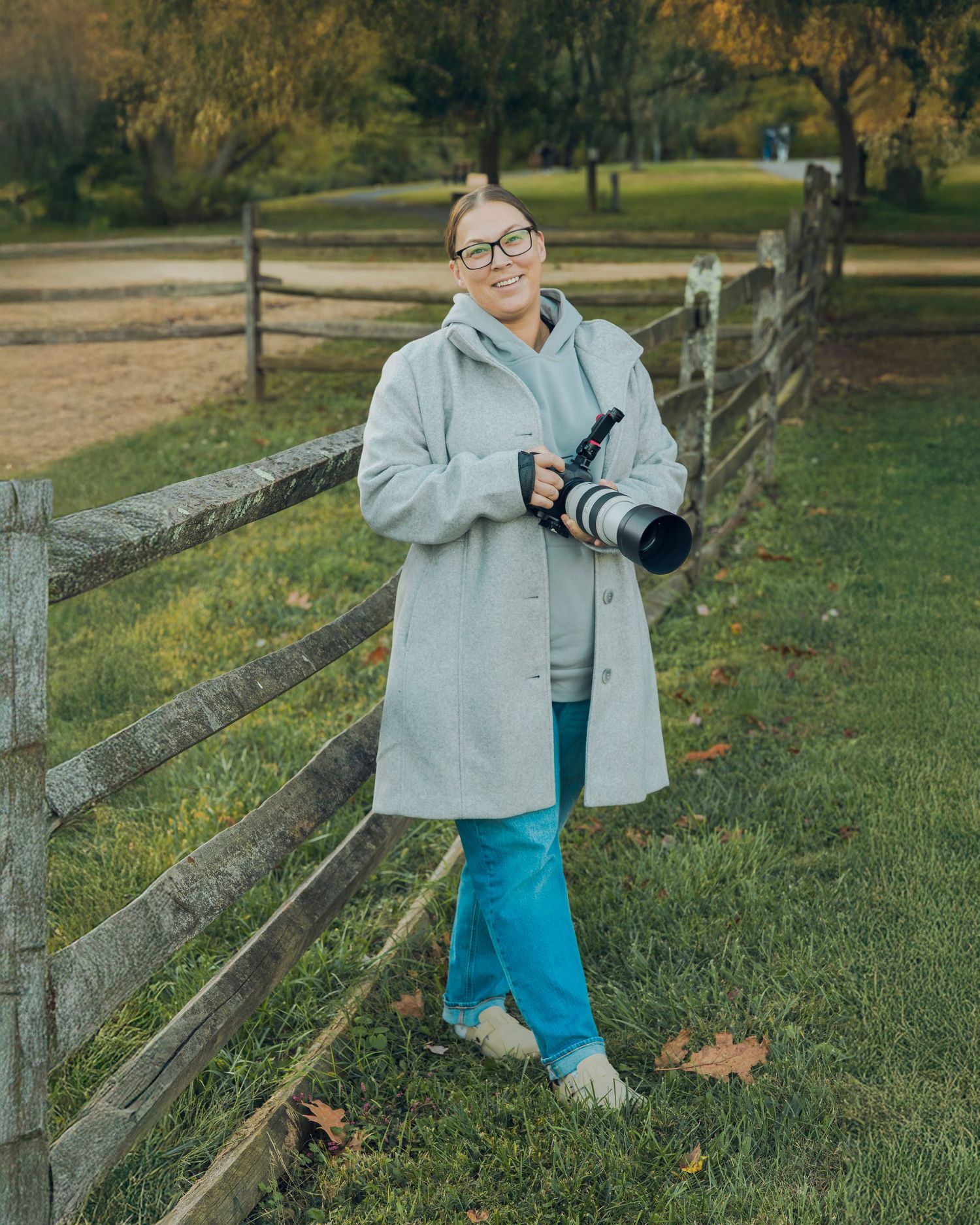 Female photographer smiling in gray coat holding camera by wooden fence in autumn park.