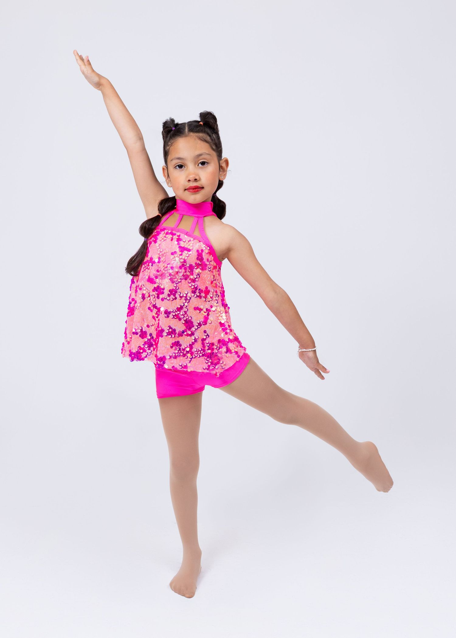 Young girl in pink sequin dance costume performs a leg extension pose in a studio photo.