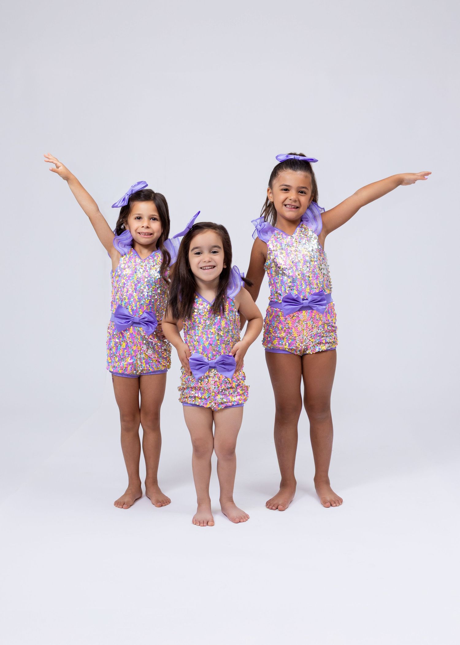 Three young girls in matching purple and gold sequin dance costumes pose together with arms raised.