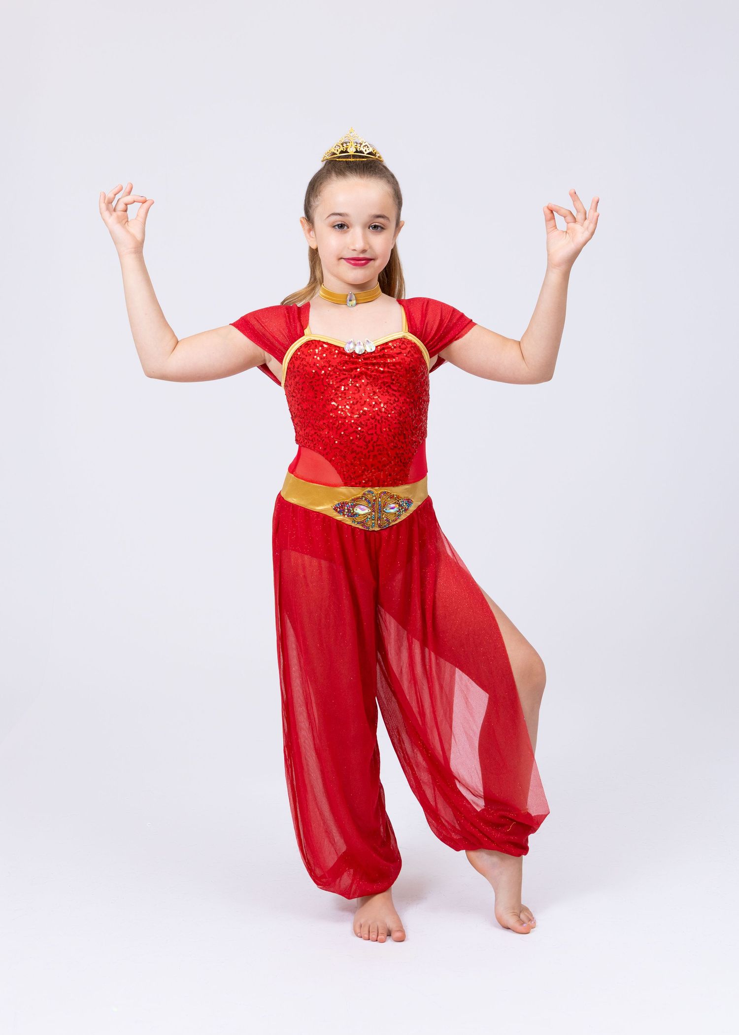 Young girl in red sequin belly dance costume with gold accessories poses gracefully with hands raised.
