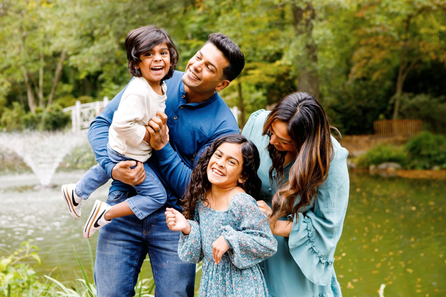 Happy South Asian family of four laughing together near a pond in a lush green park.