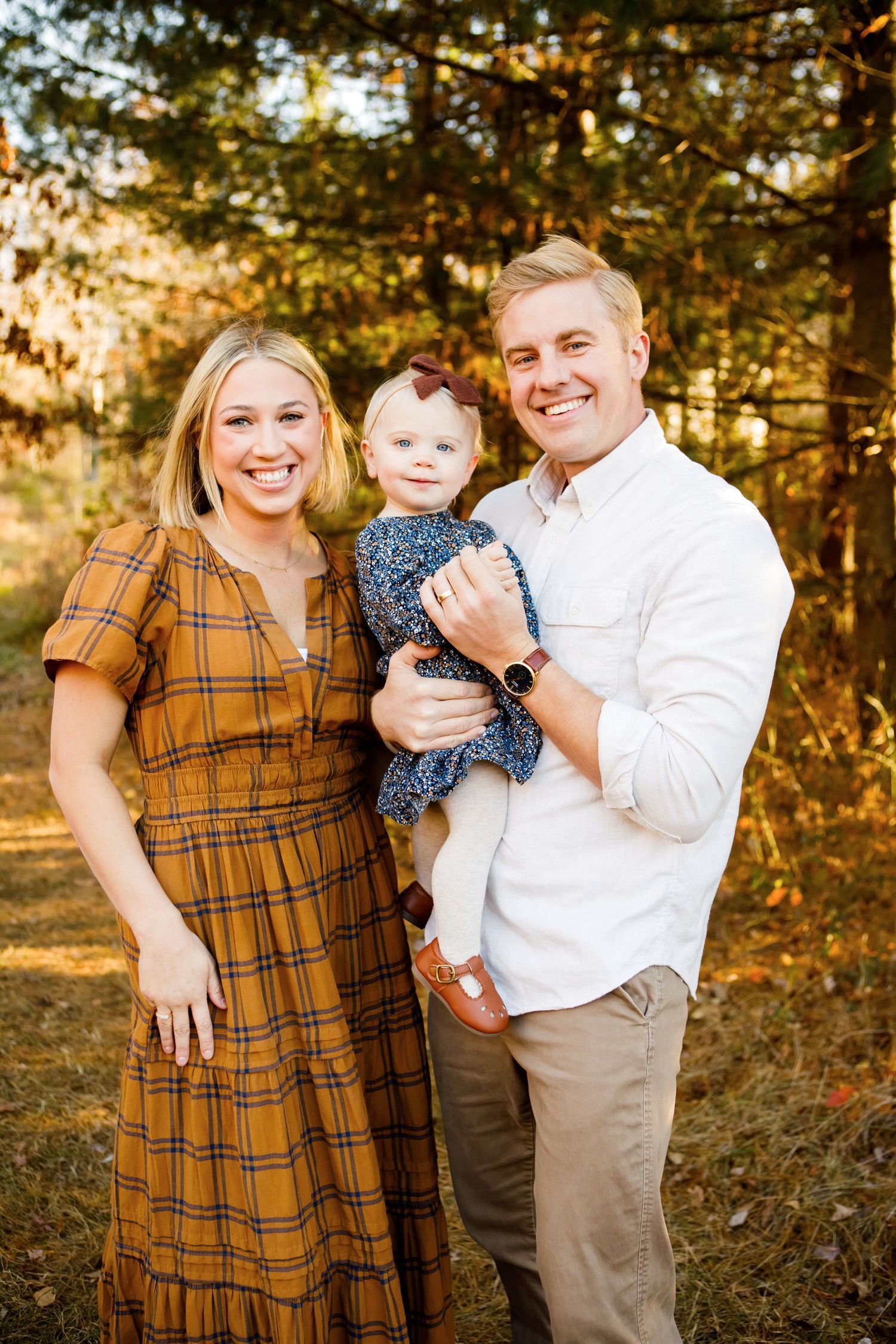 Happy family of three smiling together in a sunlit autumn pine forest setting.