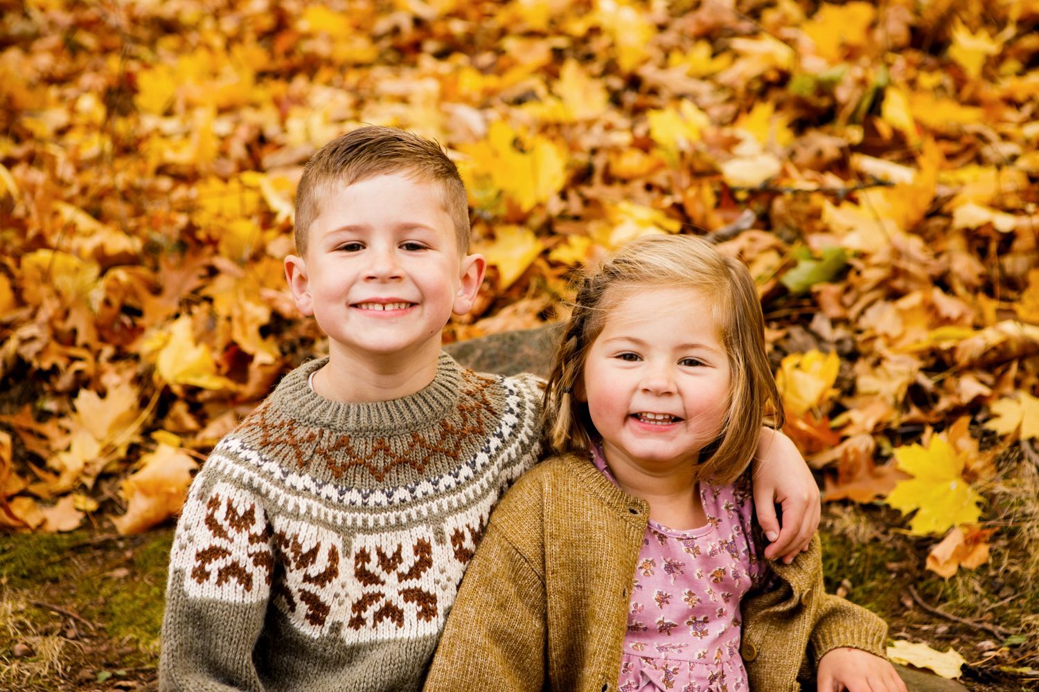 Two smiling children sitting together in a pile of golden autumn leaves.
