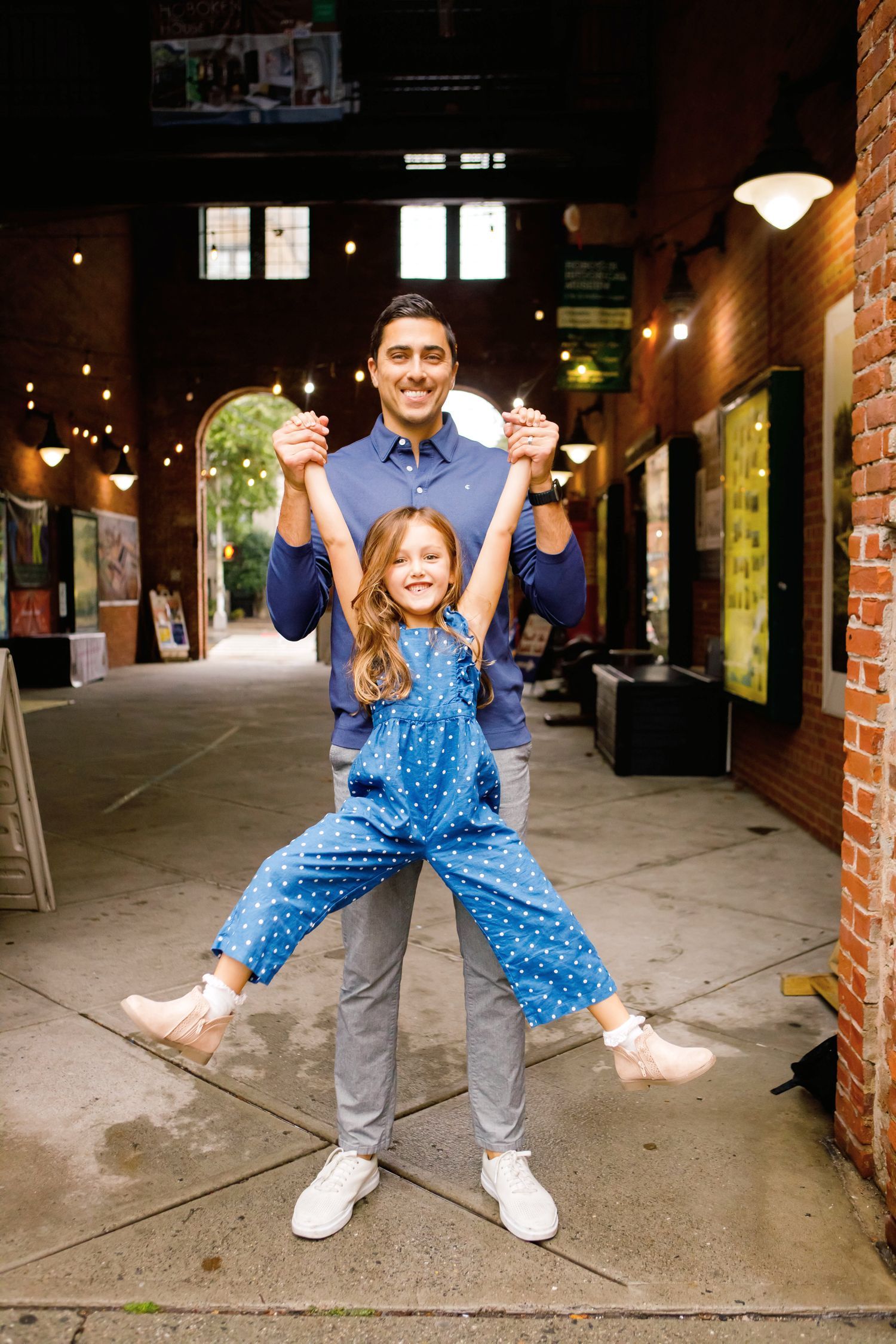 A father playfully lifting his young daughter by the hands in a brick indoor corridor.