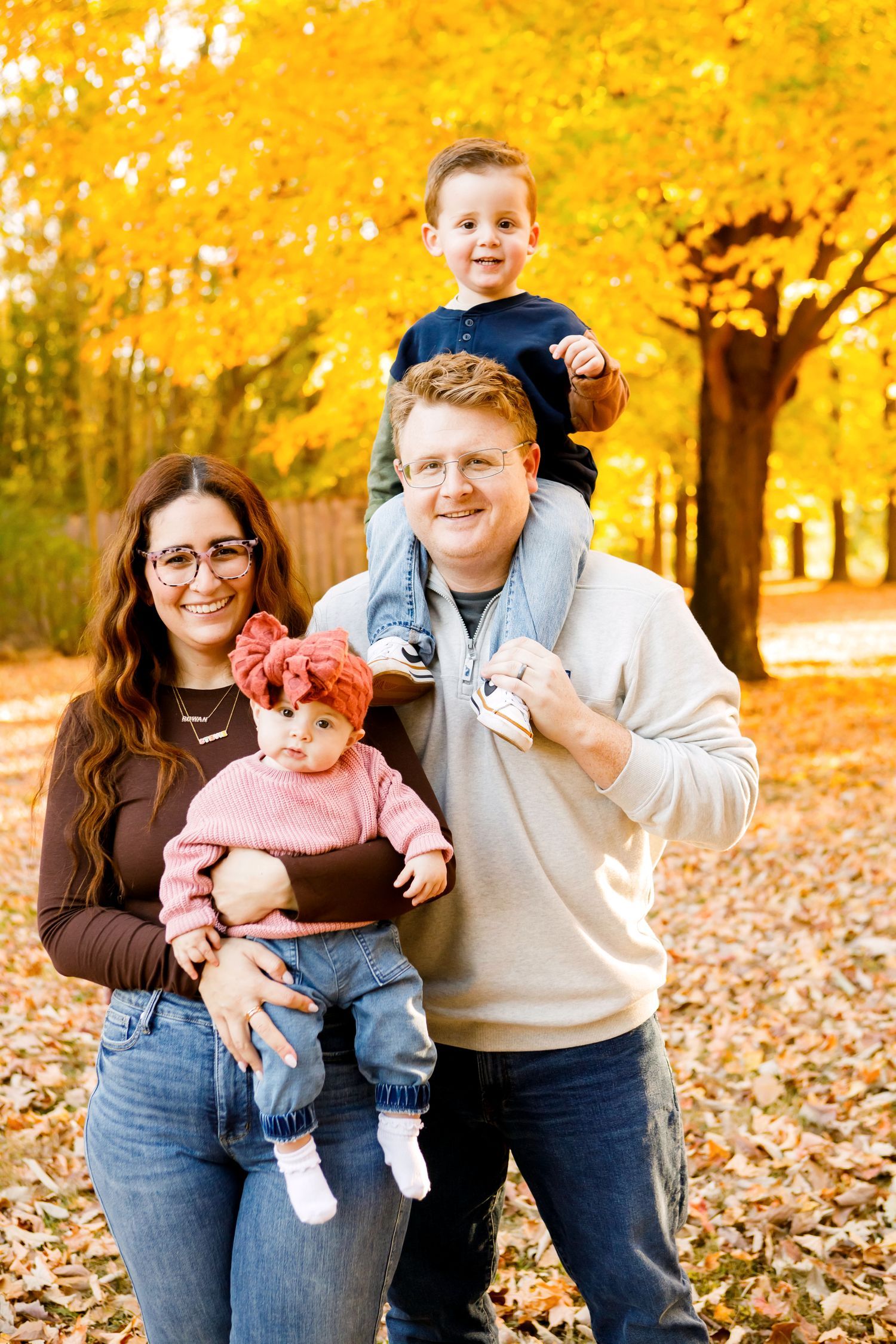 Family of four in autumn park, father carrying toddler on shoulders, mother holding baby girl.