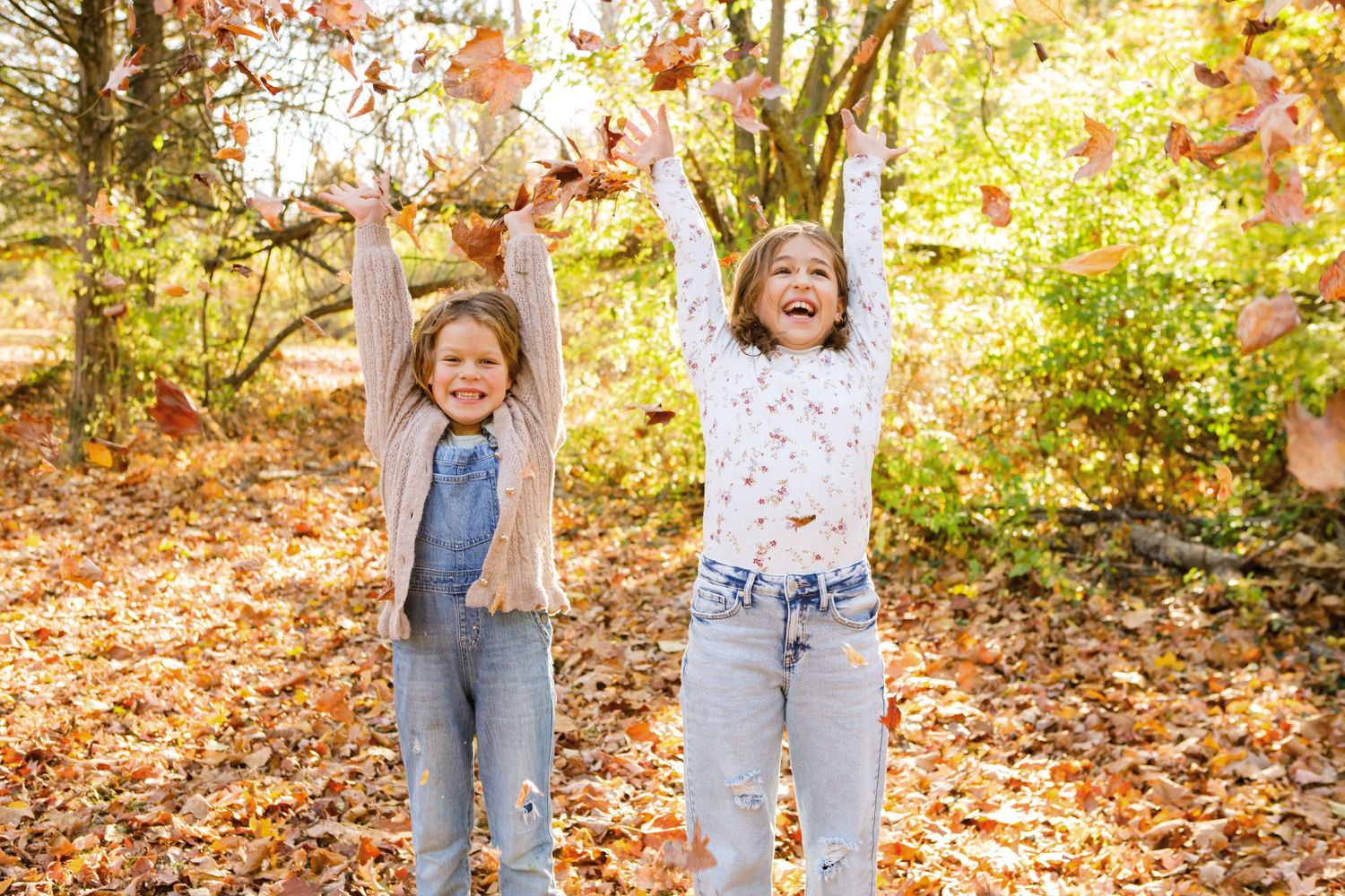 Two joyful young girls tossing autumn leaves in the air in a colorful fall forest.