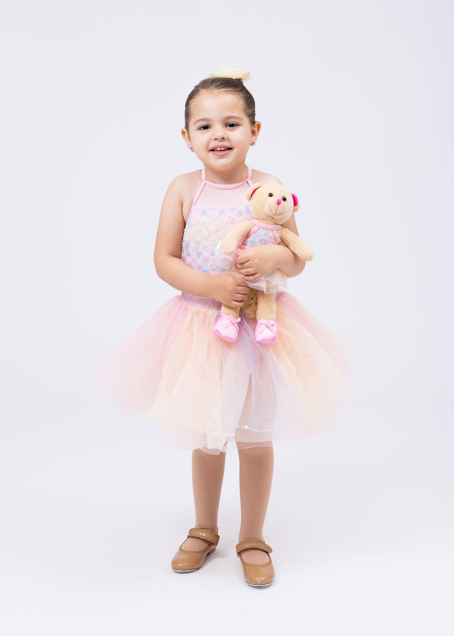 Toddler girl in pink tutu ballet dress smiles while holding a teddy bear in dance attire.