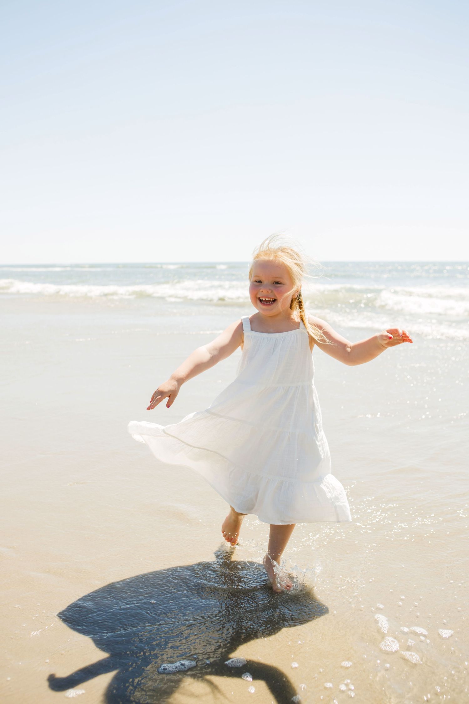 A happy little girl in a white dress playing in the ocean waves on a sunny beach day.