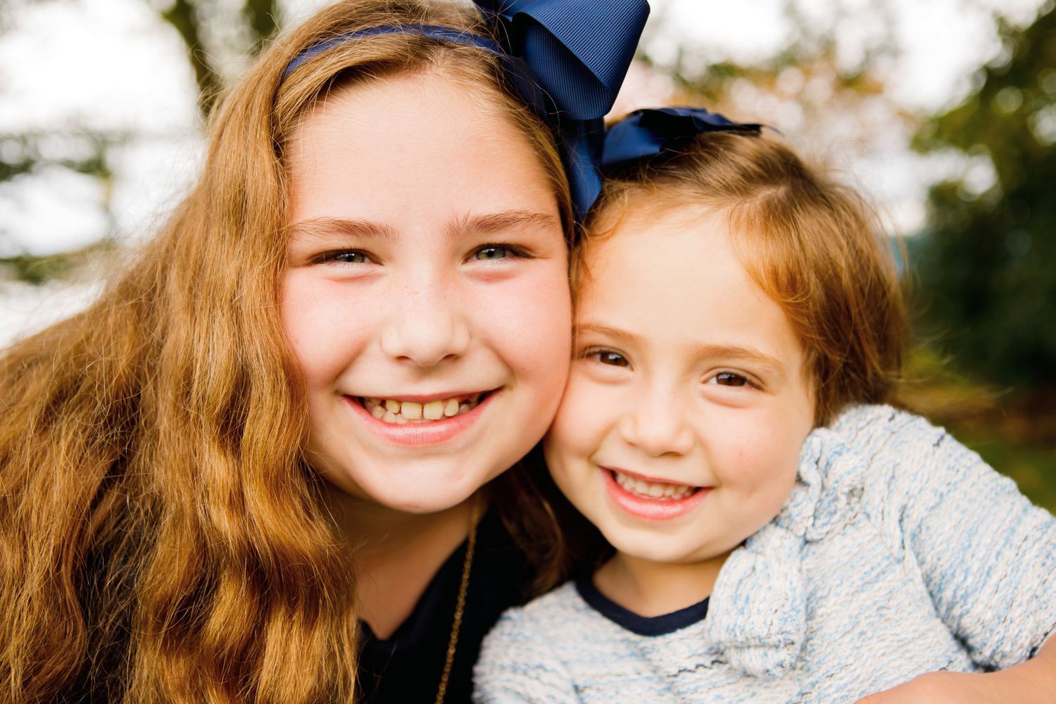 Two smiling girls pose cheek to cheek outdoors, one wearing a navy blue bow hair accessory.