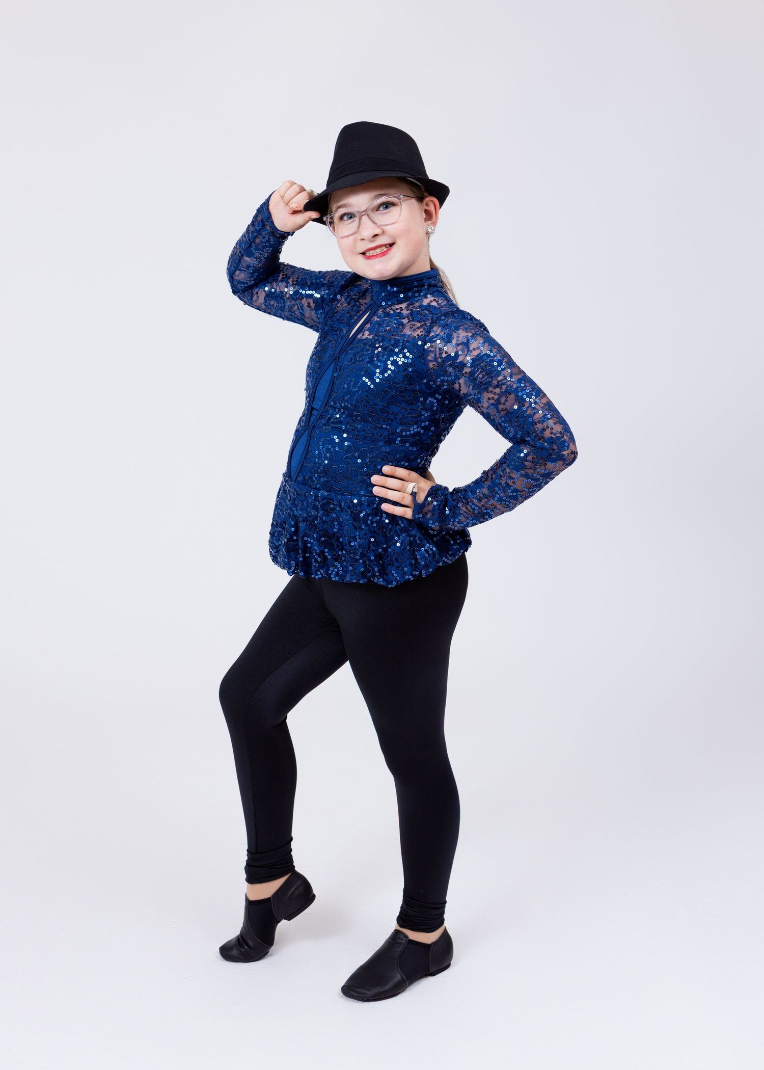Young girl in navy blue sequin lace top and black leggings poses in a black fedora hat.