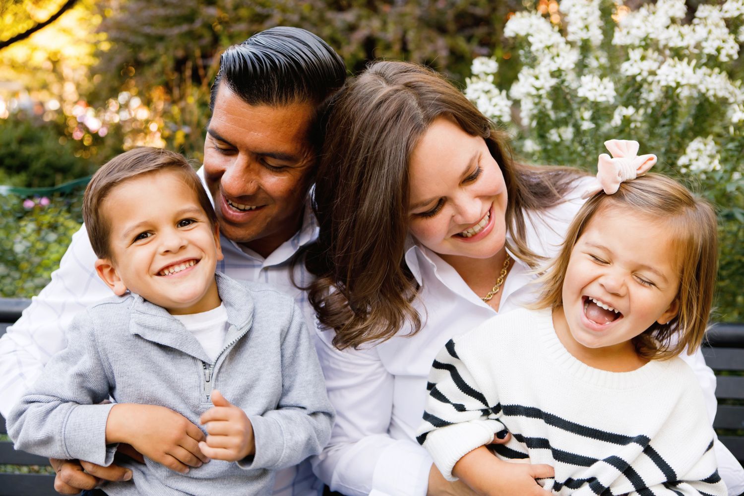 Joyful family of four laughing together outdoors with blooming white flowers in background.