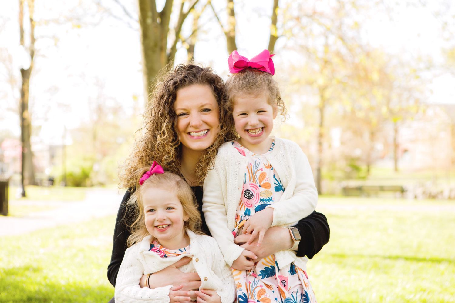 Curly-haired mother smiles holding two young daughters with pink bows in a sunny park.