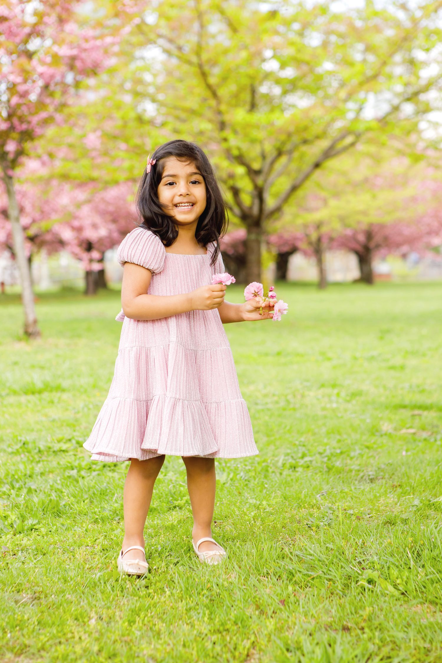 Young girl in pink dress smiles while holding cherry blossoms in a spring park.