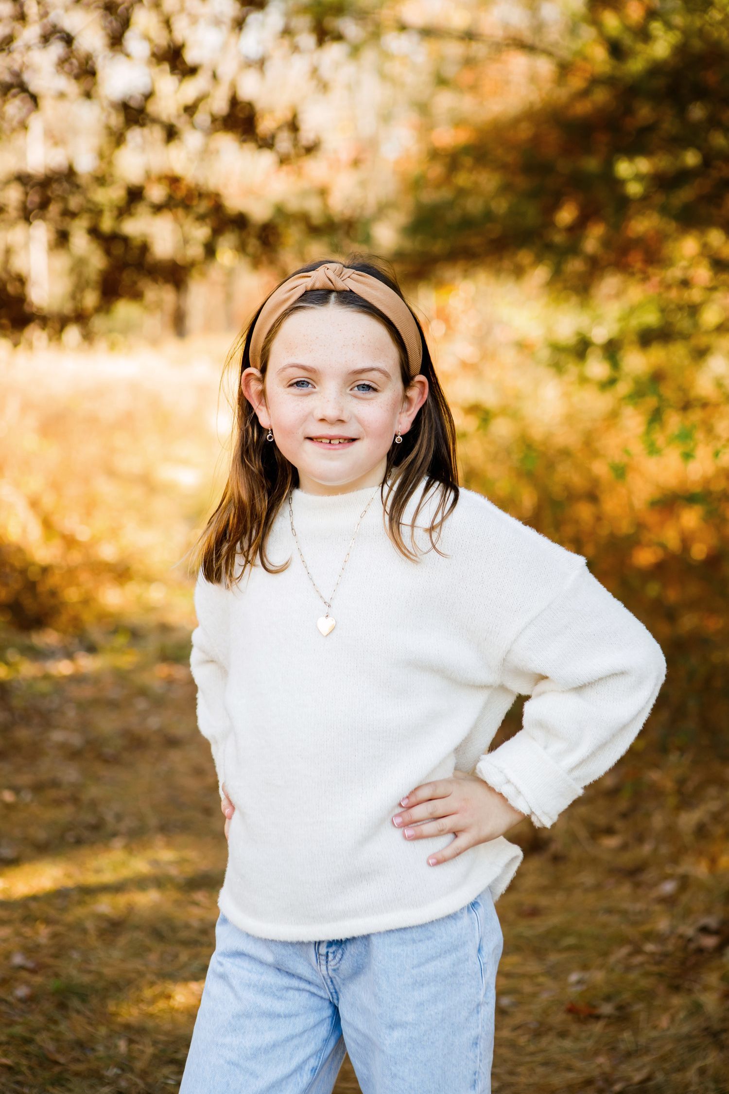 Young girl in white sweater posing confidently with hands on hips in autumn forest.