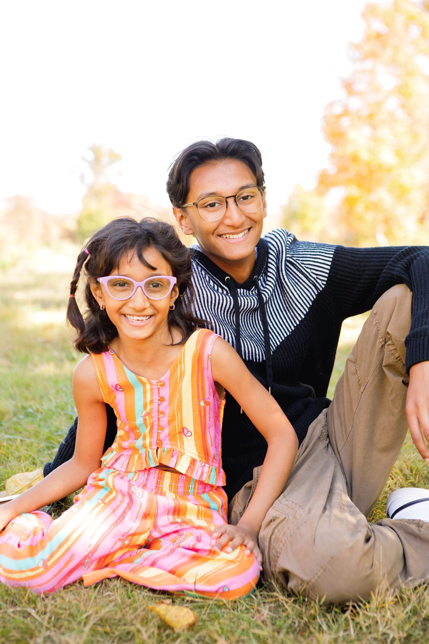 Two South Asian siblings wearing glasses sitting on grass smiling in an outdoor park.