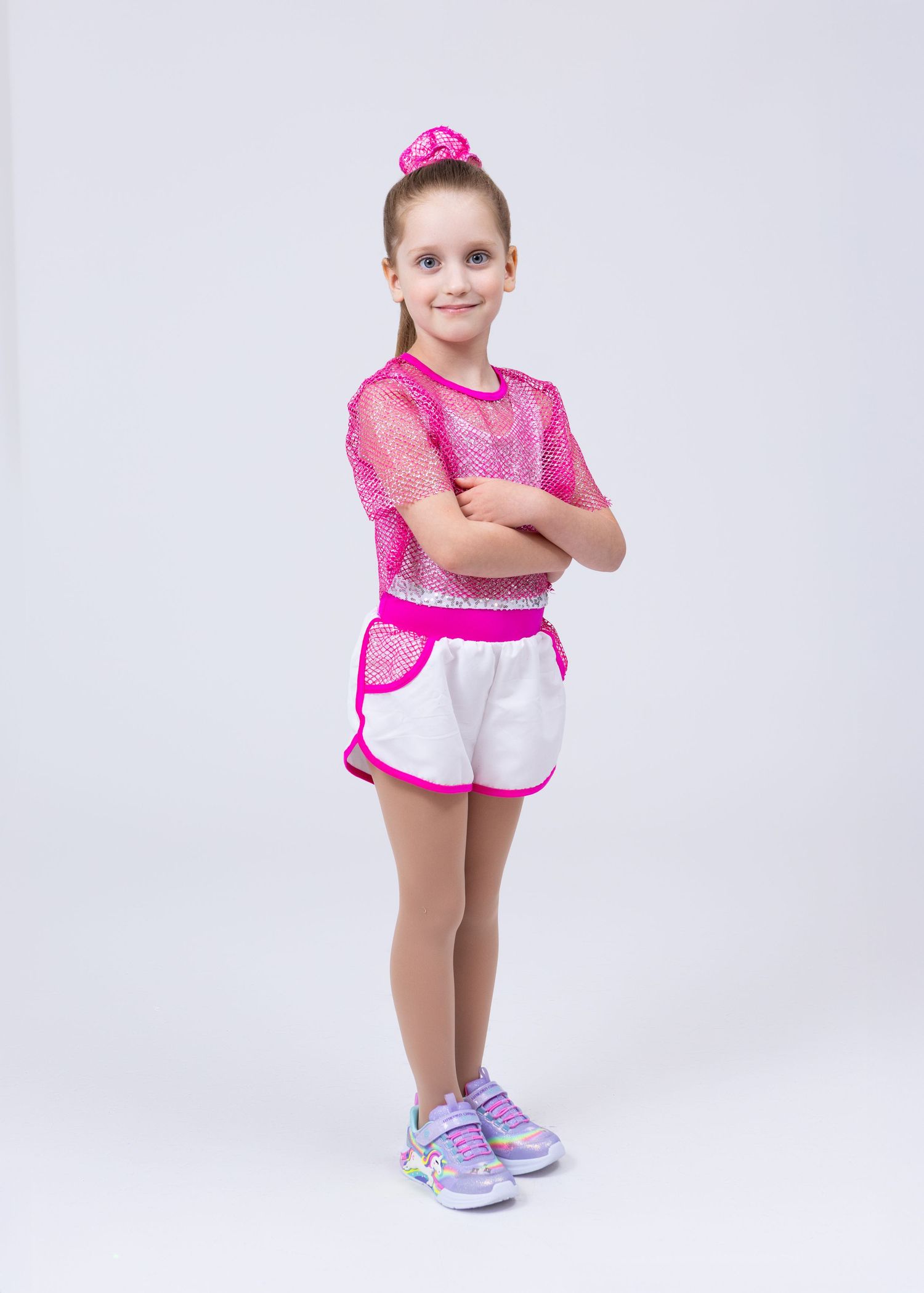 A young girl in a pink mesh top and white shorts with pink trim stands with arms crossed, smiling.