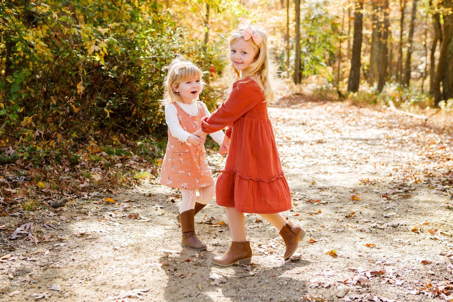 Two little sisters in orange and pink dresses hold hands dancing on an autumn forest path.