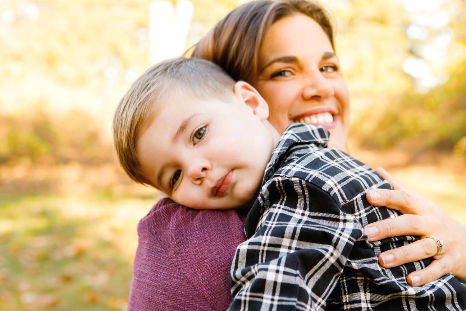 A smiling mother hugging her young son outdoors in a sunny autumn park setting.