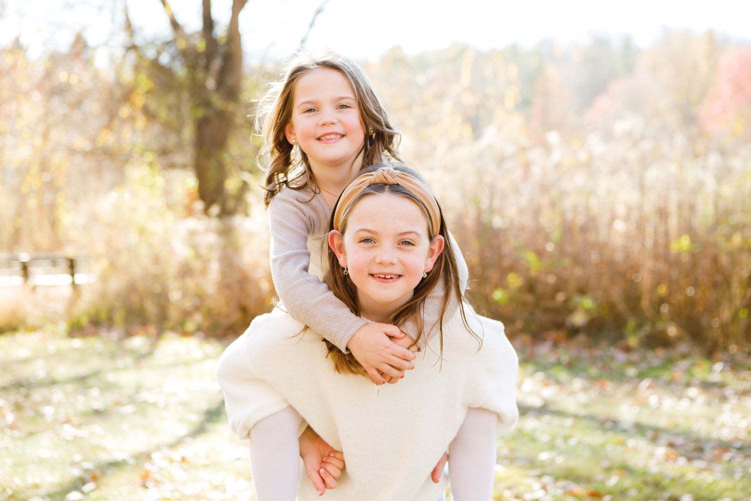 Two girls laughing, one giving a piggyback ride in a sunny autumn outdoor setting.