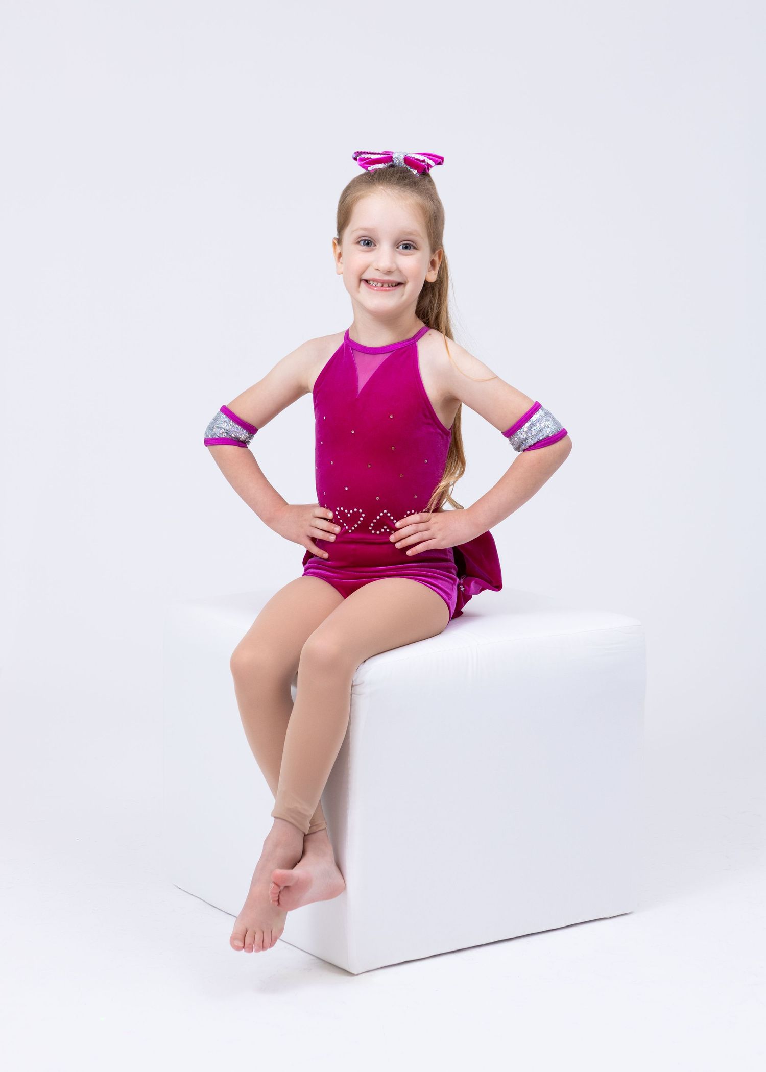 A young girl in a pink sequined dance costume sits on a white cube, smiling at the camera.