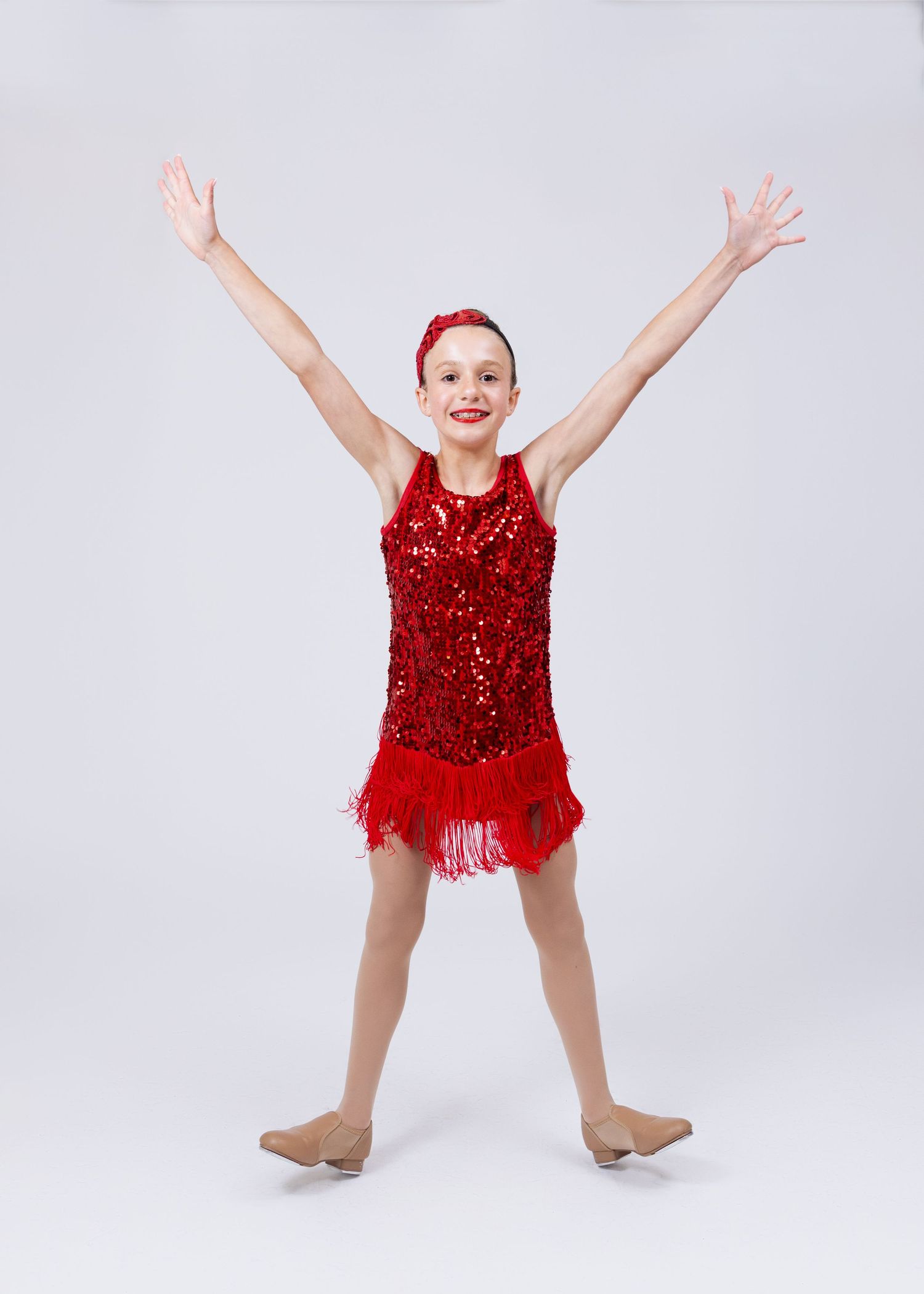 Young girl in red sequin fringe dance dress performs with arms raised wide in a victory pose.