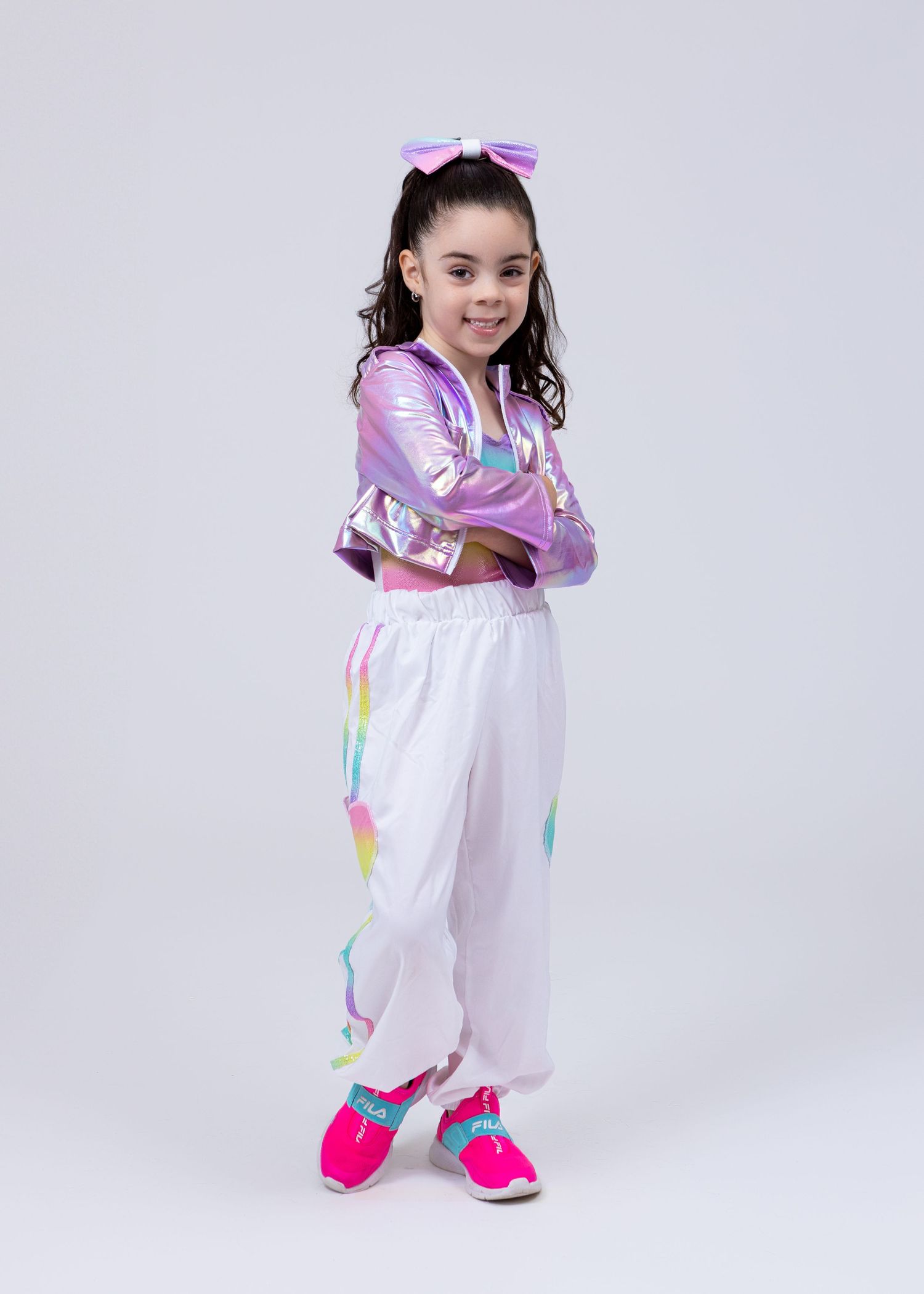Young girl in iridescent purple jacket and white pants with pink bow poses with arms crossed.